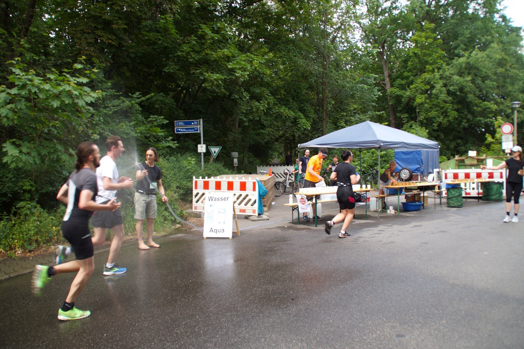 Mehrere Personen joggen an einem kleinen Stand vorbei und werden mit einem Wasserschlauch nassgespritzt