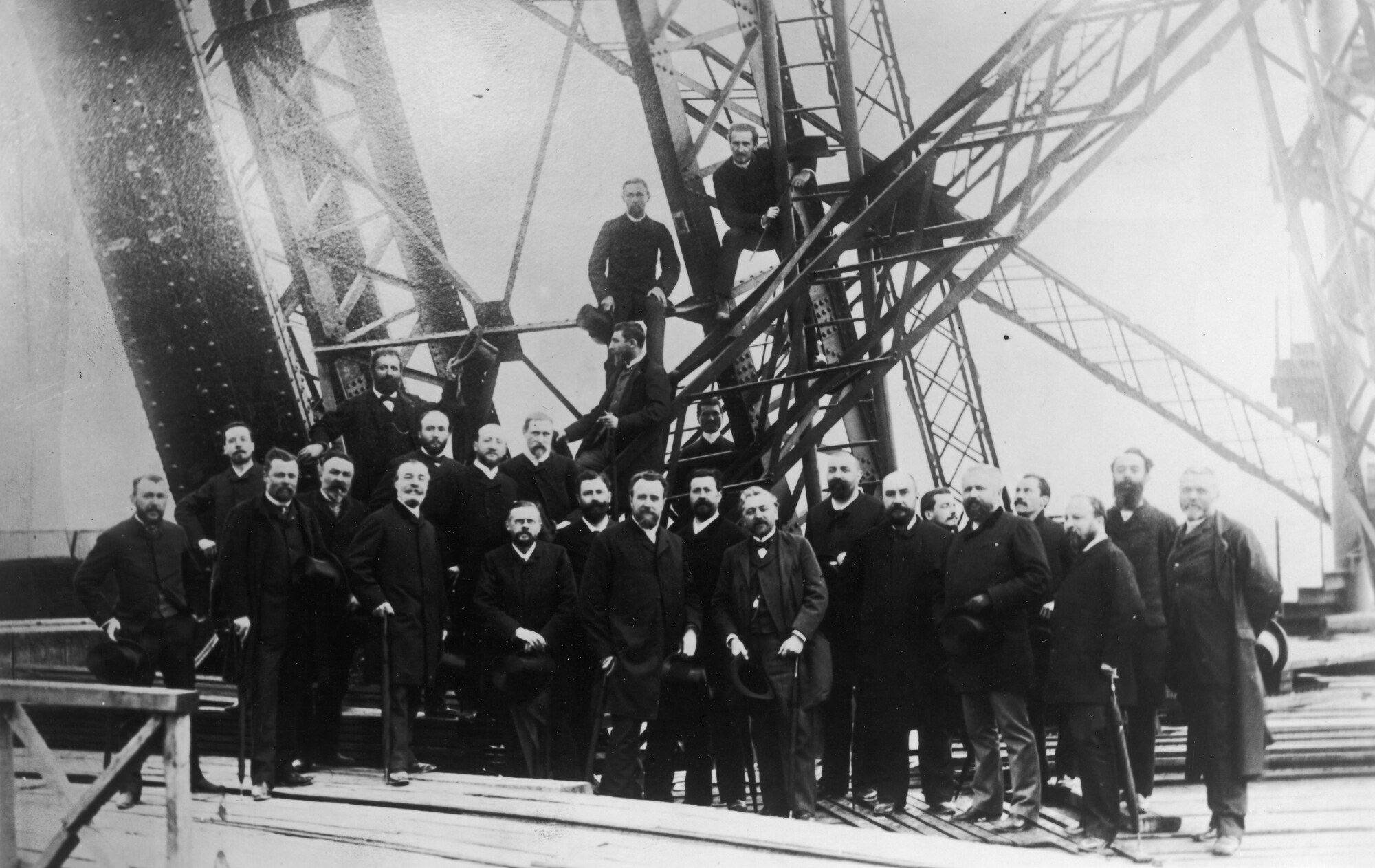 Schwarzweißaufnahme von Gustave Eiffel mit einer Gruppe Männer vor einer Stahlkonstruktion beim Bau des Eiffelturms.