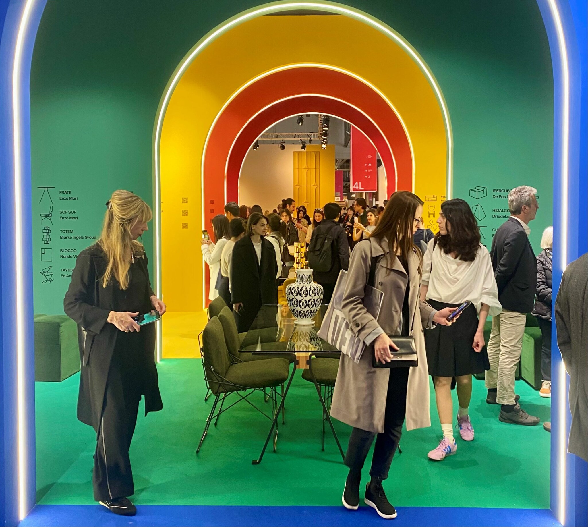 Menschen auf dem Messestand von Driade mit Torbögen in Gelb, Rot und Grün. Menschen auf dem Messestand von Driade mit Torbögen in Gelb, Rot und Grün.