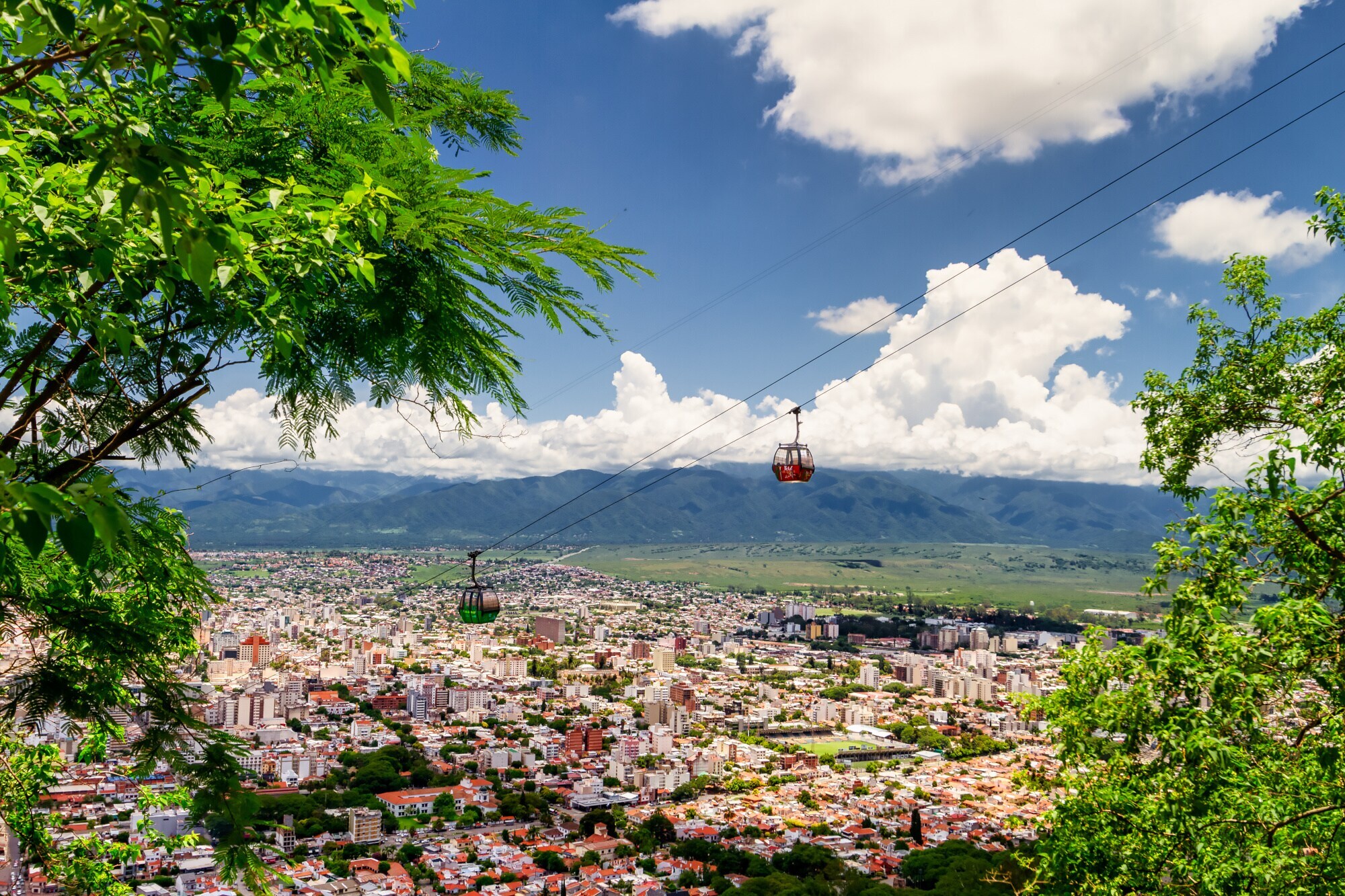 Panoramablick auf eine Seilbahn mit zwei Gondeln, die über der Stadt Salta einen Berg hinauffahren, im Hintergrund hügeliges Umland Panoramablick auf eine Seilbahn mit zwei Gondeln, die über der Stadt Salta einen Berg hinauffahren, im Hintergrund hügeliges Umland