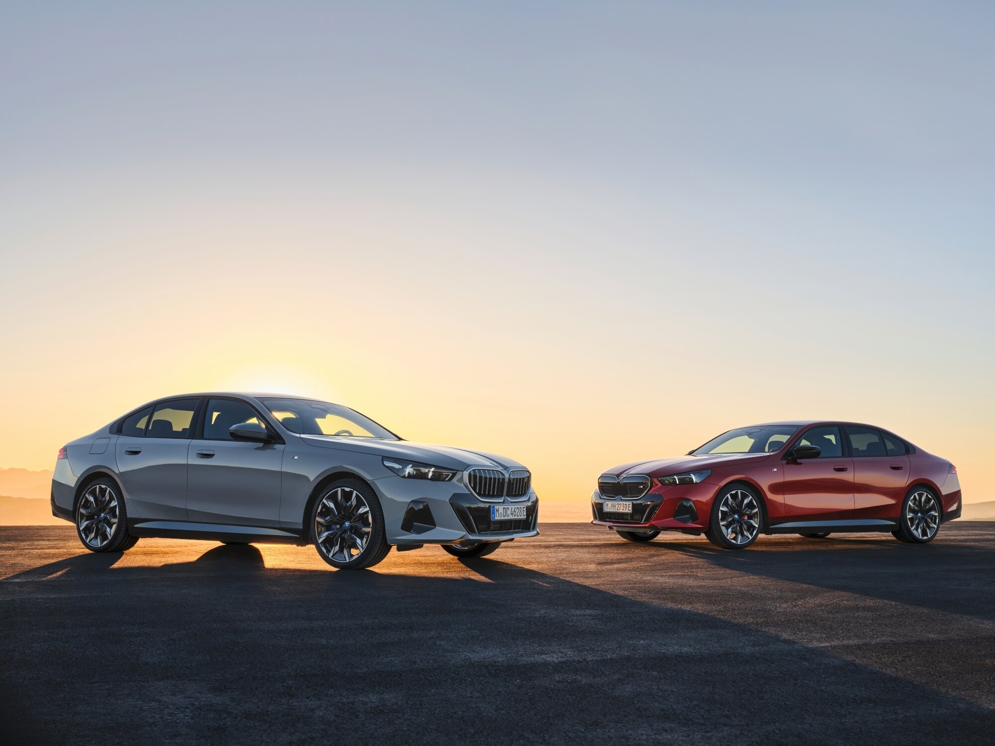 Zwei BMW i5, einmal in Silber, einmal in Rot, die von hinten von der Sonne angestrahlt werden. Zwei BMW i5, einmal in Silber, einmal in Rot, die von hinten von der Sonne angestrahlt werden.