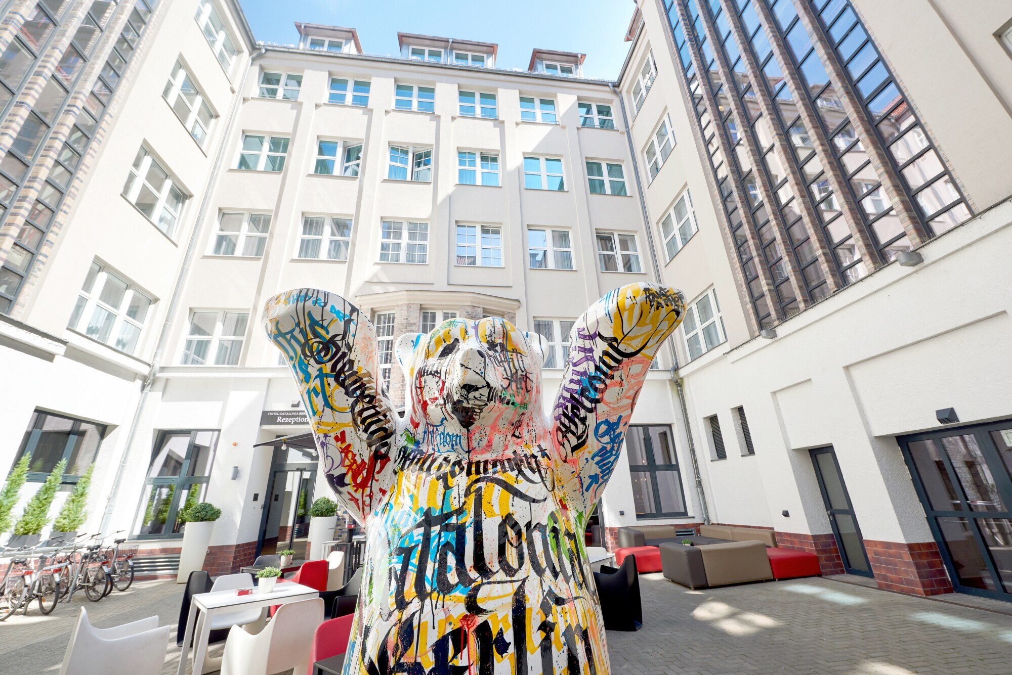 Eine mit Graffiti bunt bemalte Statue eines Berliner Bären steht im sonnigen Innenhof eines mehrstöckigen weißen Hotels.