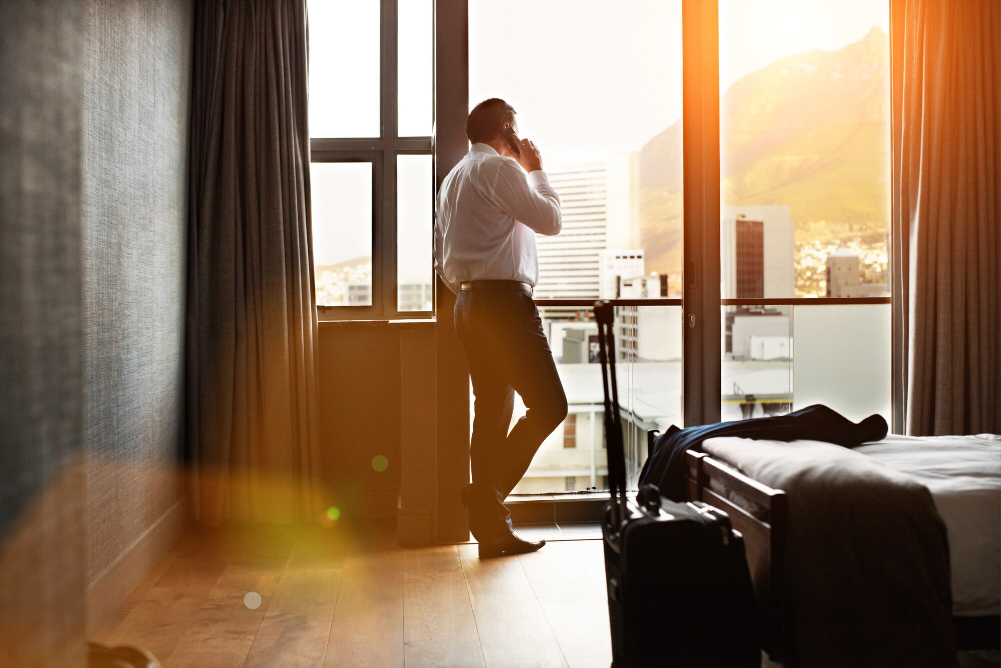 Rückansicht eines Geschäftsmannes, der durch ein Hotelzimmerfenster schaut und auf einem Mobiltelefon spricht Rückansicht eines Geschäftsmannes, der durch ein Hotelzimmerfenster schaut und auf einem Mobiltelefon spricht