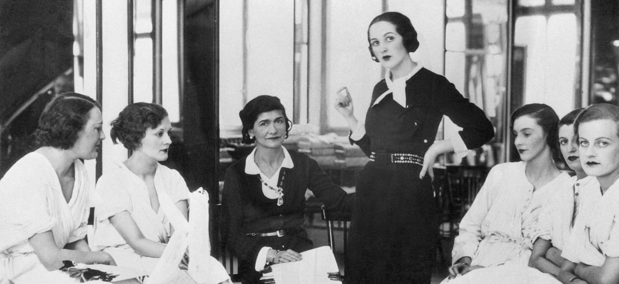 Schwarzweißfoto von Coco Chanel in einem schwarzen Kostüm, umgeben von weiblichen Models in ihrem Modeatelier