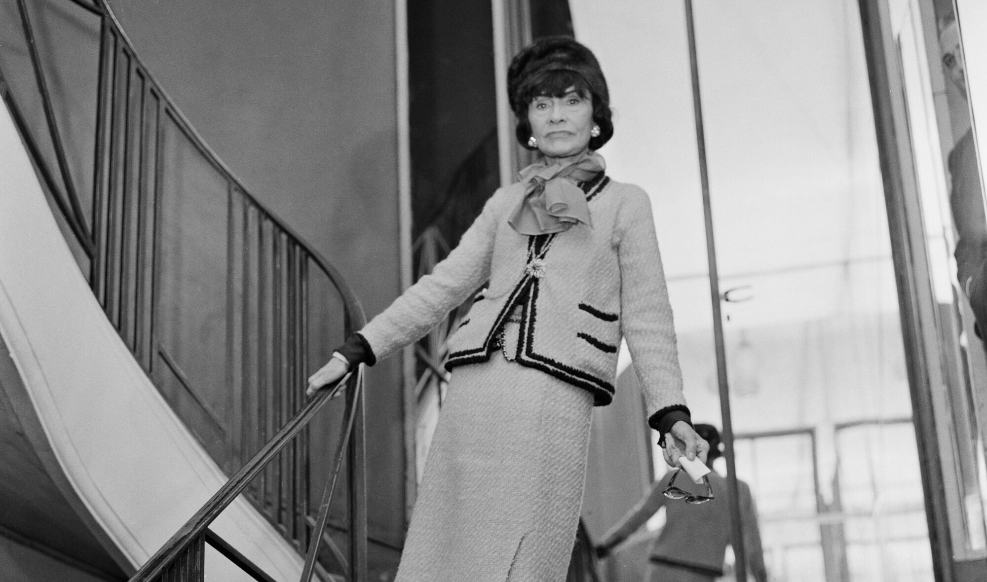 Schwarzweißaufnahme von Coco Chanel in einem Tweed-Kostüm auf einer Treppe vor einer Spiegelwand Schwarzweißaufnahme von Coco Chanel in einem Tweed-Kostüm auf einer Treppe vor einer Spiegelwand