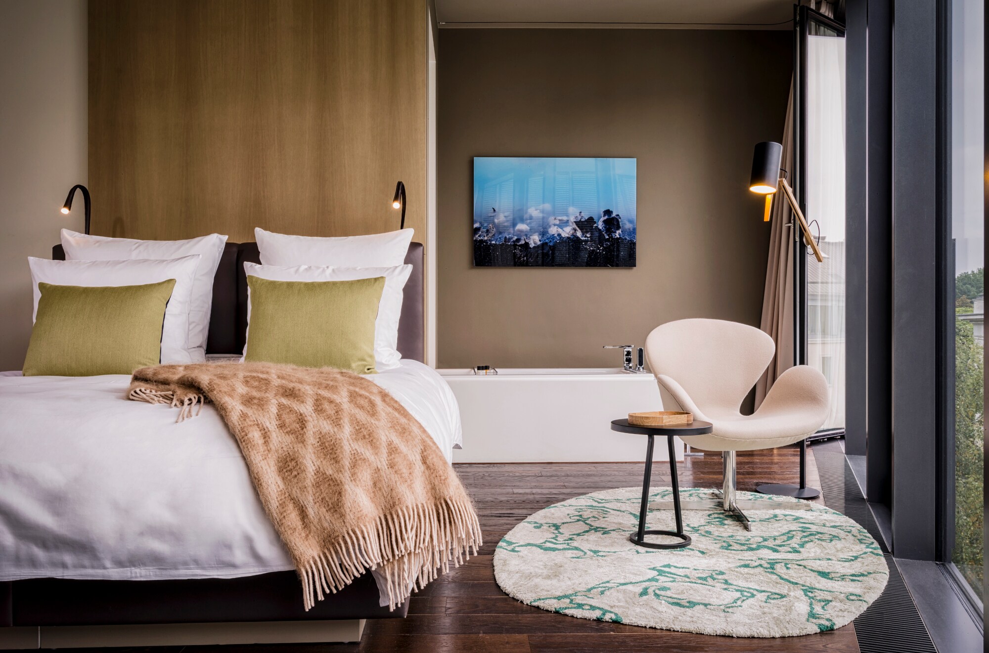Blick auf ein Zimmer im Berliner Buisinesshotel So Berlin Das Stue mit Bett, Sessel und Beistelltisch Modernes, stilvolles Hotelzimmer mit bodentiefen Fenstern.