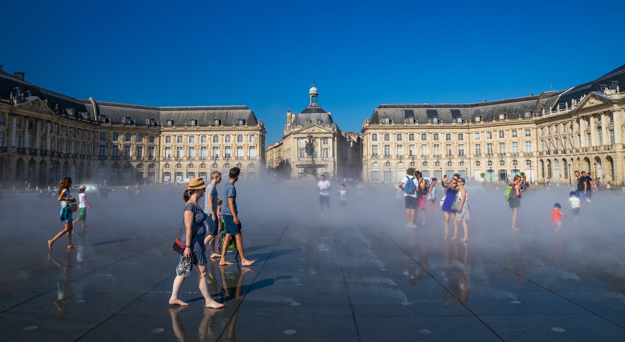 Personen in Sommerbekleidung laufen über einen Wasserspiegel auf einem großen Platz vor neoklassizistischen Gebäuden in Bordeaux Personen in Sommerbekleidung laufen über einen Wasserspiegel auf einem großen Platz vor neoklassizistischen Gebäuden in Bordeaux