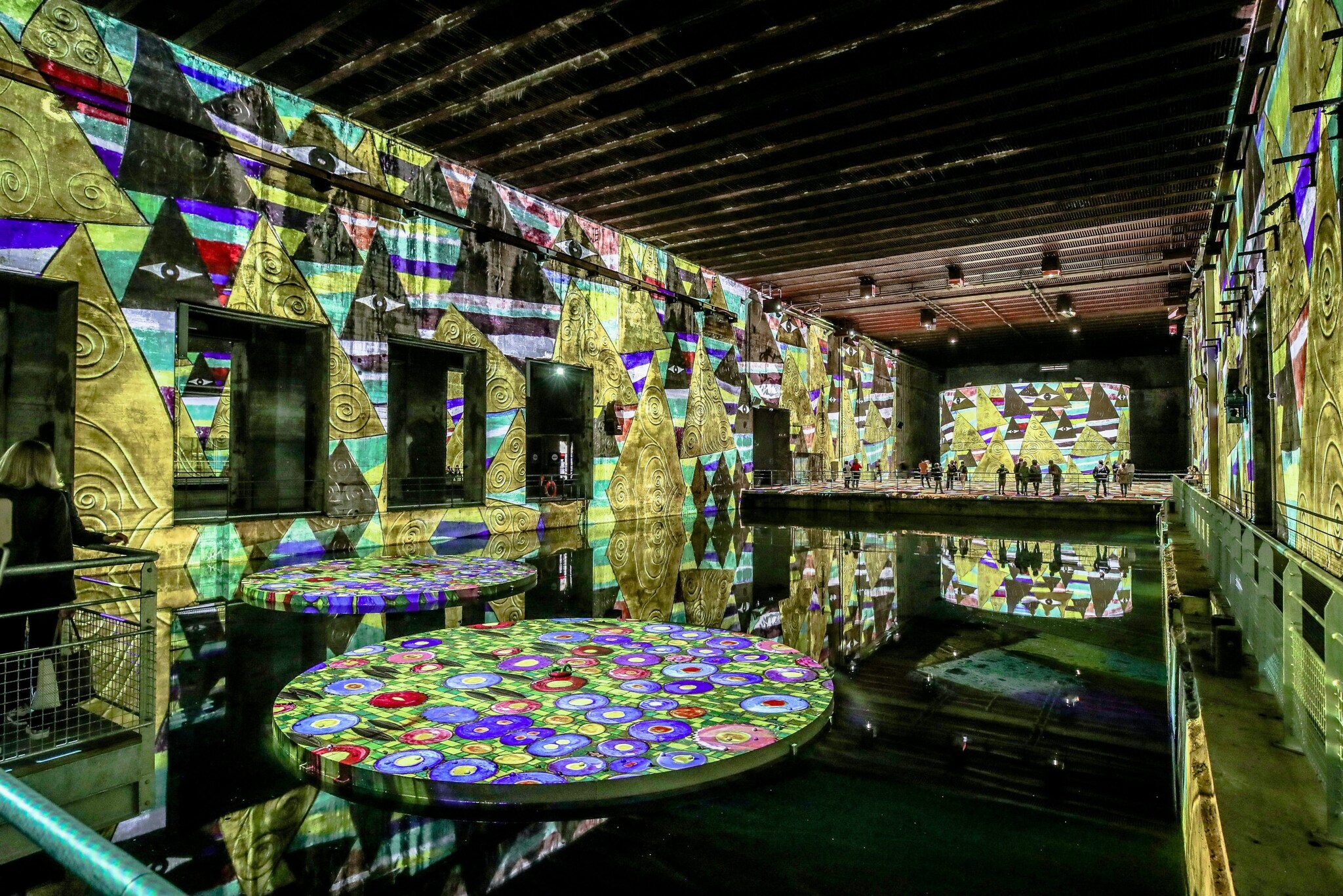 Besucher einer digitalen Lichtausstellung in einer alten Fabrikhalle mit an die Wände projizierten Kunstwerken des Malers Gustav Klimt