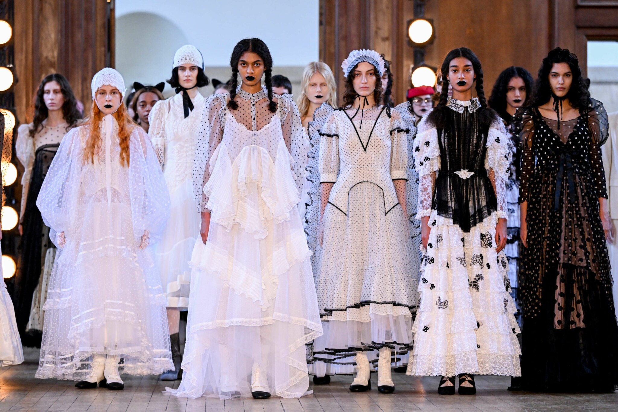 Models in langen, verspielten Kleidern in schwarz und weiß stehen nebeneinander auf dem Laufsteg einer Modenschau