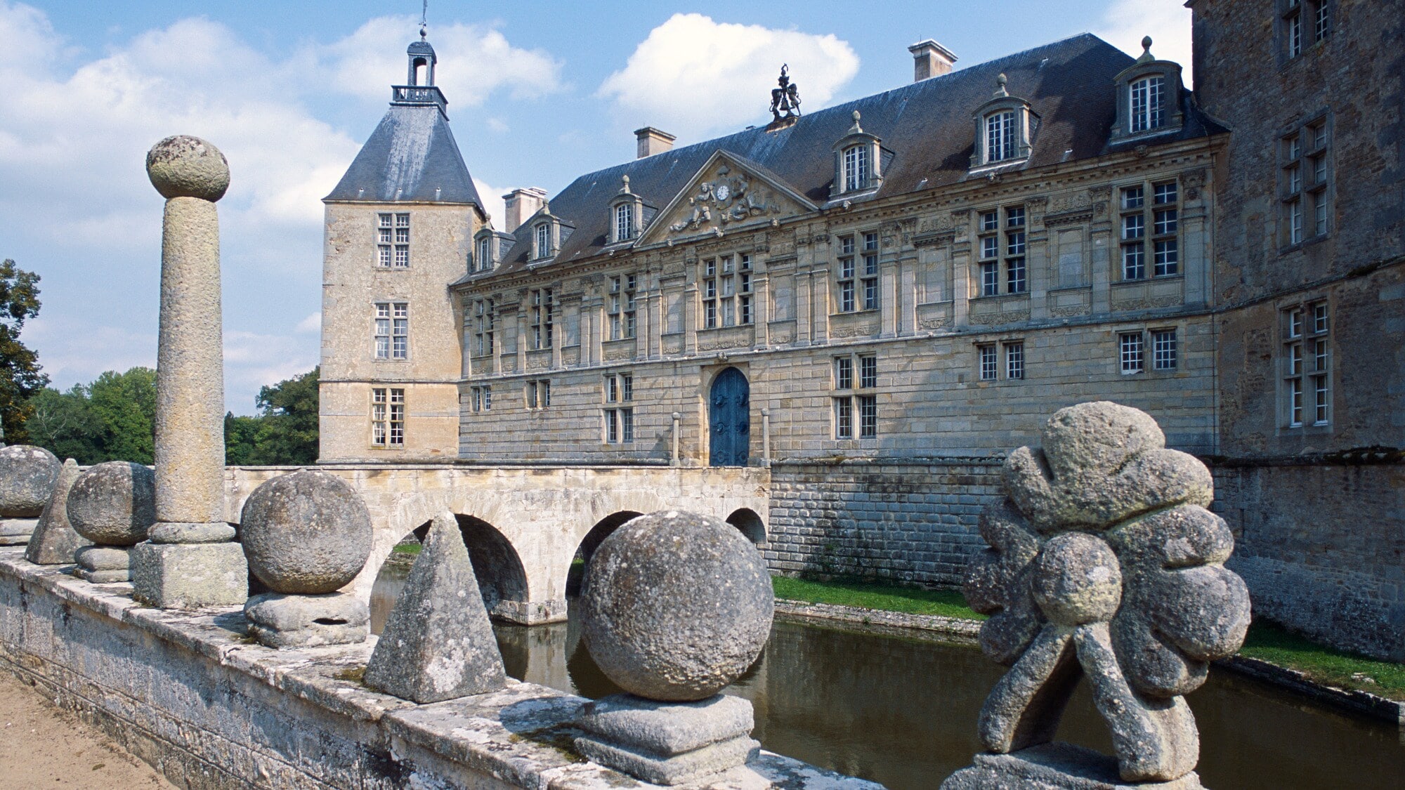 Ein französisches Wasserschloss mit Brücke und Steinskulpturen auf einer Mauer im Vordergrund Ein französisches Wasserschloss mit Brücke und Steinskulpturen auf einer Mauer im Vordergrund