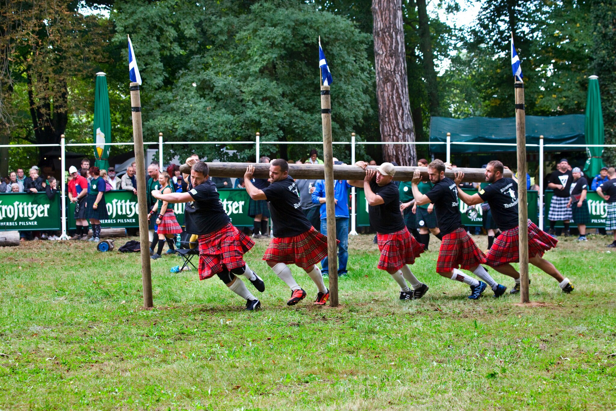 Fünf Personen in Kilts tragen einen Baumstamm und rennen damit zwischen senkrechten Holzpflöcken hindurch. Fünf Personen in Kilts tragen einen Baumstamm und rennen damit zwischen senkrechten Holzpflöcken hindurch.