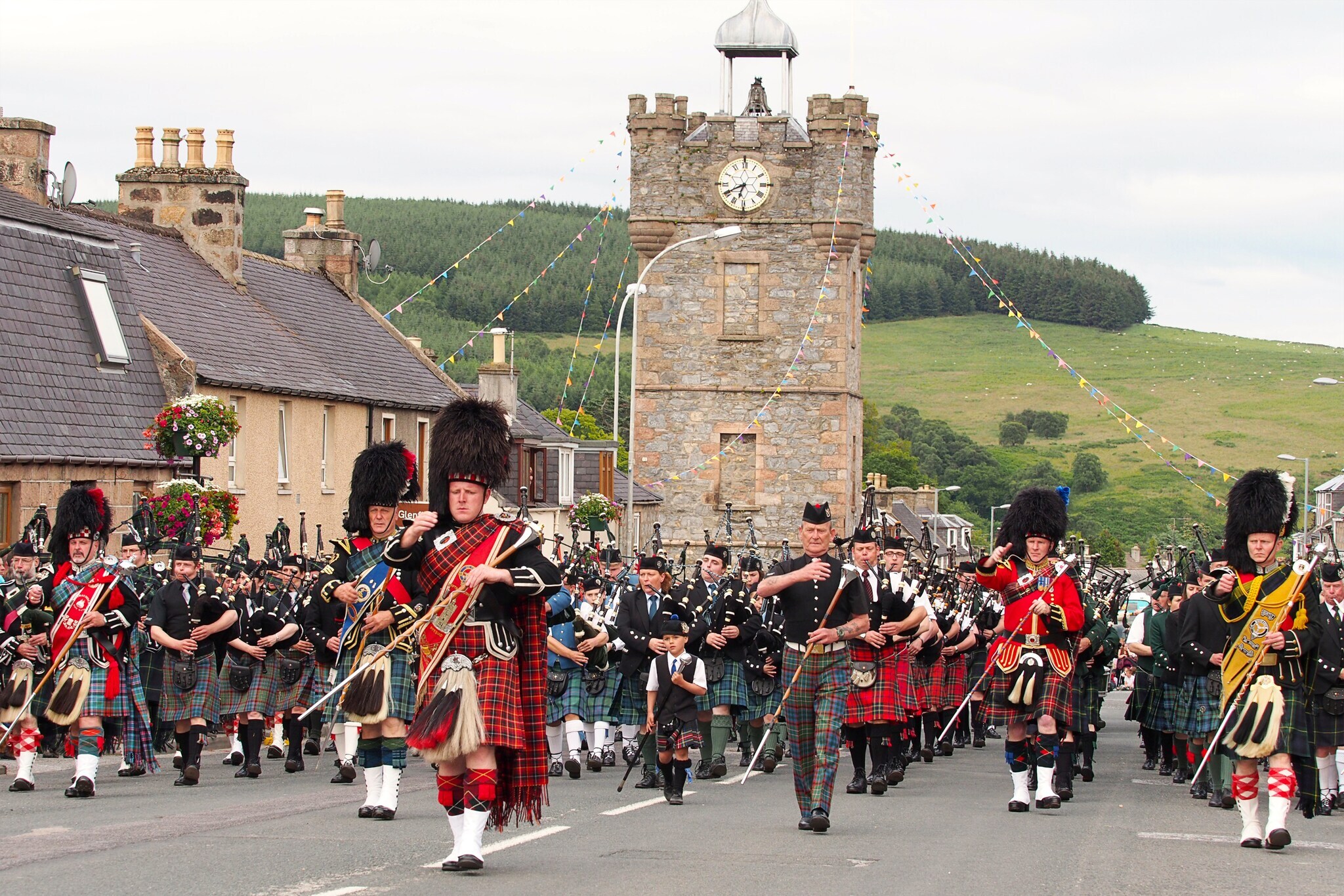 Diverse Menschen in traditionellen schottischen Outfits, die eine Straße entlanglaufen und Instrumente spielen.