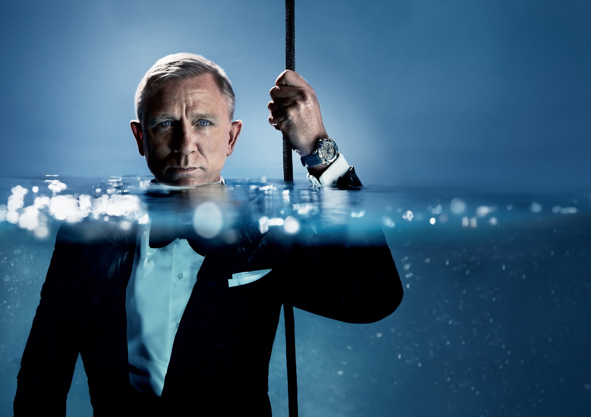 Porträt von Daniel Craig im schwarzen Smoking im Wasser, das ihm bis zum Hals steht; an seiner hochgehaltenen Hand trägt er eine Taucheruhr der Marke OMEGA Porträt von Daniel Craig im schwarzen Smoking im Wasser, das ihm bis zum Hals steht; an seiner hochgehaltenen Hand trägt er eine Taucheruhr der Marke OMEGA