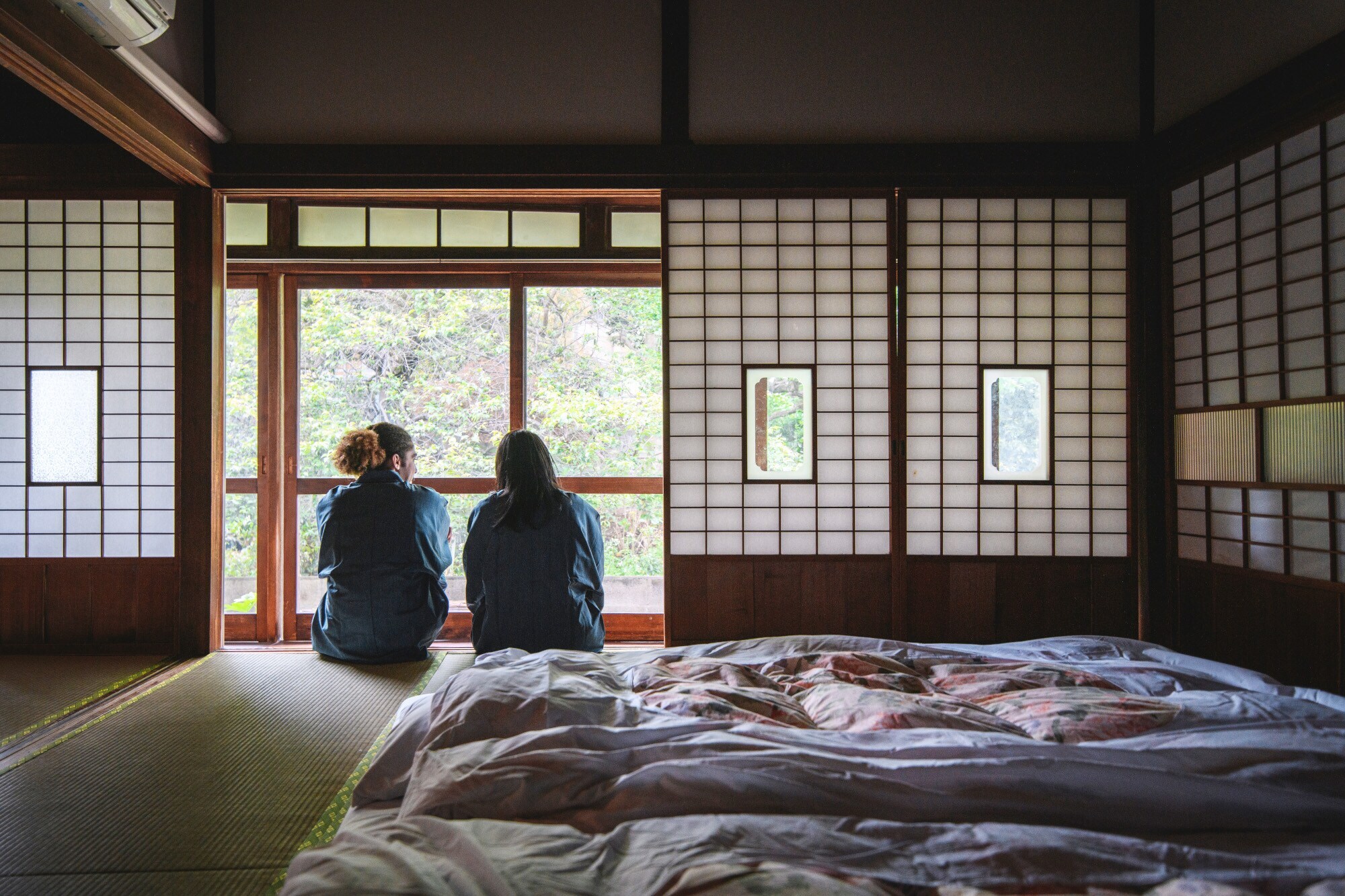 Rückansicht zweier Personen, die neben einem Futonbett in einer japanischen Herberge sitzen und aus dem Fenster in einen Garten schauen