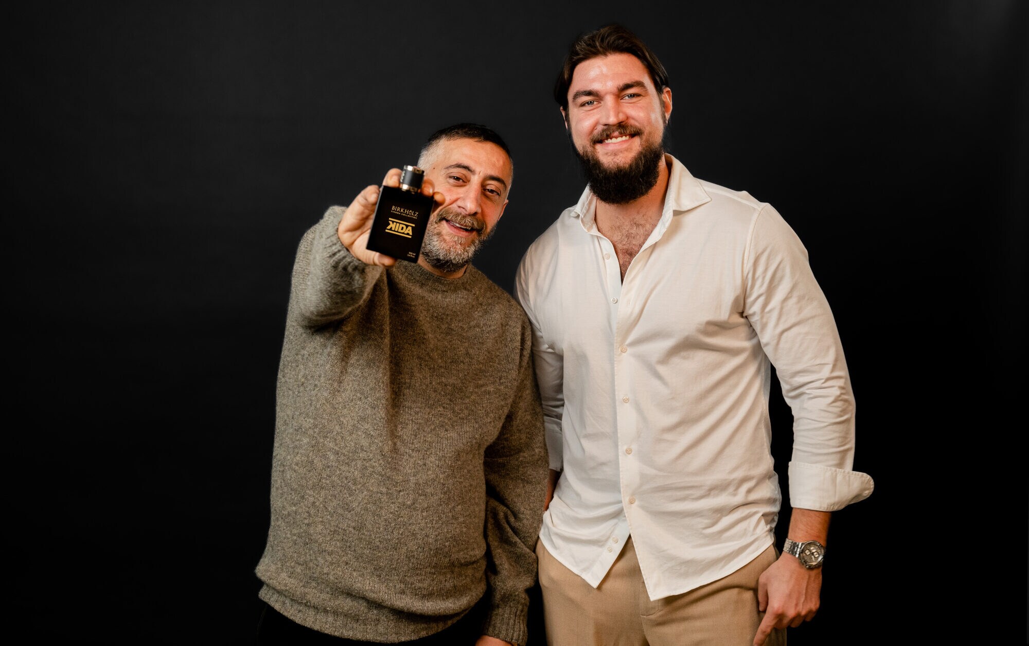 Kida Khodr Ramadan und Philip Birkholz stehen Schulter an Schulter vor einem schwarzen Hintergrund. Kida Khodr Ramadan hält den Flakon des Parfüms KIDA in die Kamera