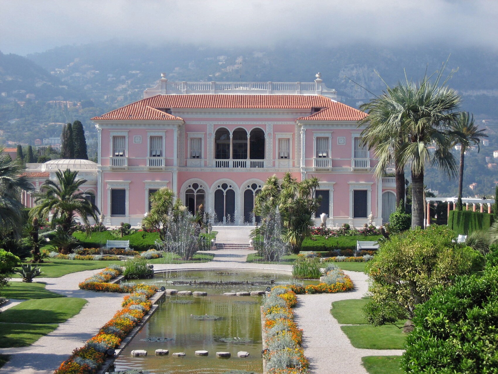 Blick auf die rosafarbene Fassade der Villa Ephrussi de Rothschild, vor der sich ein Teich erstreckt und die in einer gepflegten Parkanlage steht Blick auf die rosafarbene Fassade der Villa Ephrussi de Rothschild, vor der sich ein Teich erstreckt und die in einer gepflegten Parkanlage steht