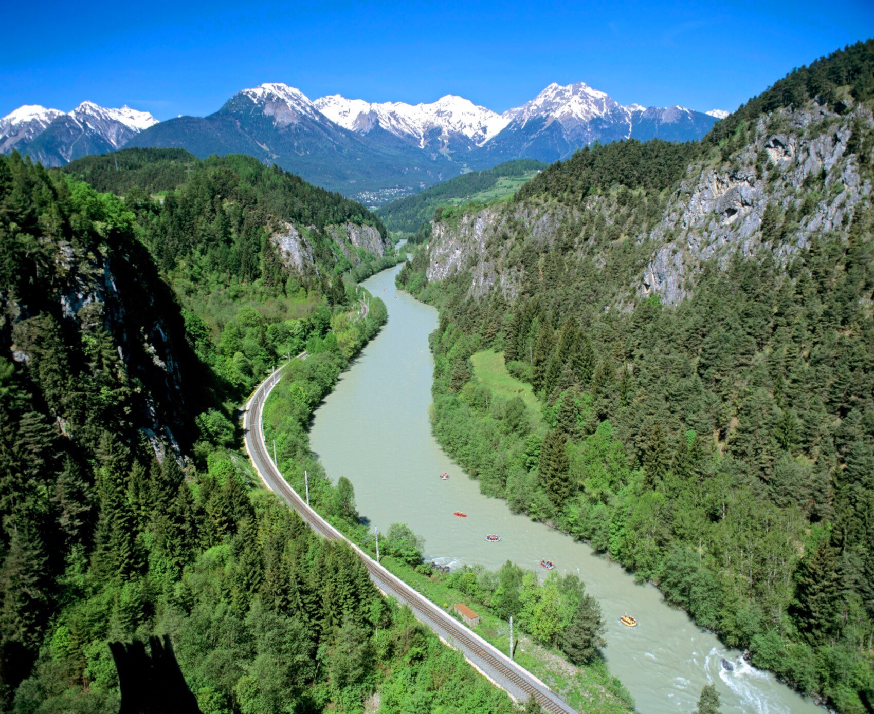 Alpenpanorama mit Waldgebiet und einem breiten Fluss, auf dem Raftingboote fahren