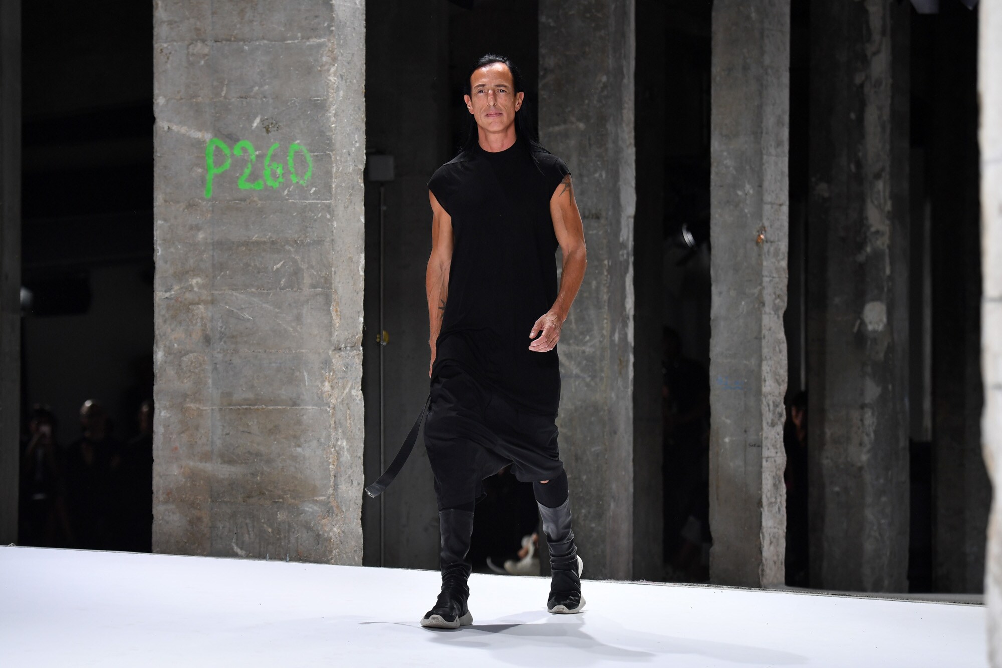 Designer Rick Owens in einem schwarzen, lässigen Kleid läuft in einer Industriehalle über einen Lauftsteg