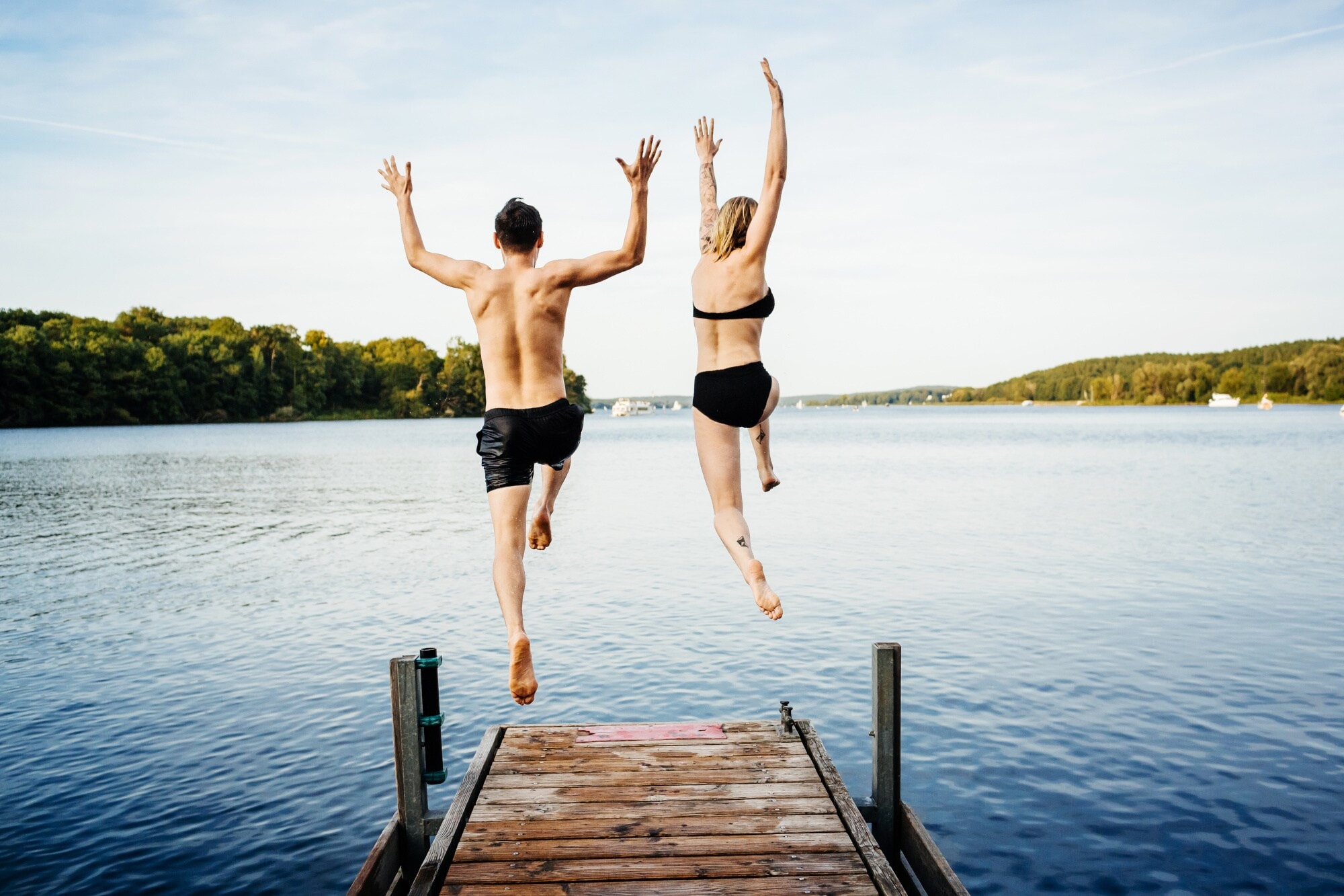 Rückansicht eines Paares in Badekleidung, das von einem Steg in einen See springt Rückansicht eines Paares in Badekleidung, das von einem Steg in einen See springt