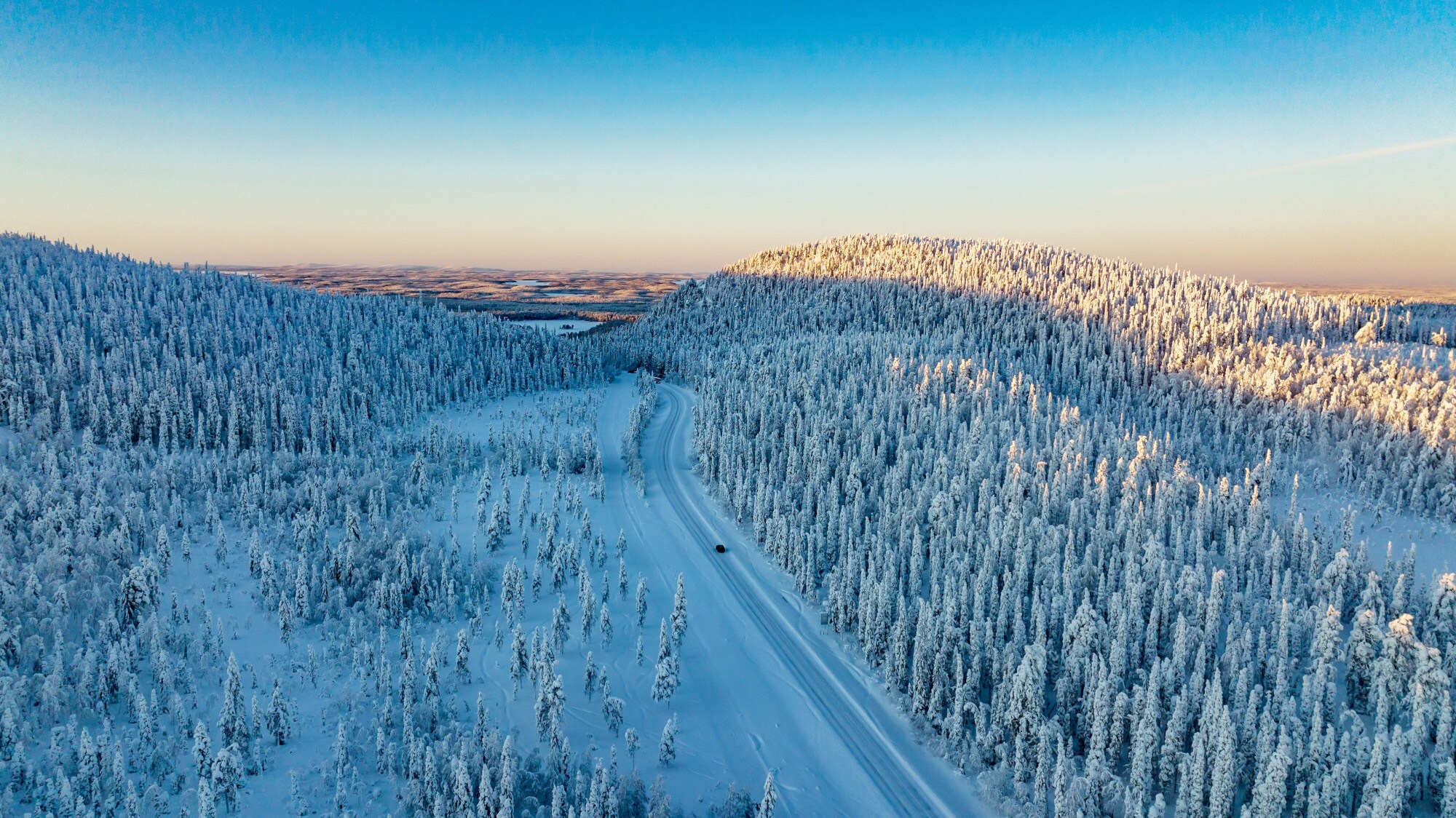 Luftaufnahme einer weiten Winterlandschaft mit schneebedeckten Wäldern, durch die ein einzelnes Auto über eine Straße durch den Schnee in der Abenddämmerung fährt Luftaufnahme einer weiten Winterlandschaft mit schneebedeckten Wäldern, durch die ein einzelnes Auto über eine Straße durch den Schnee in der Abenddämmerung fährt