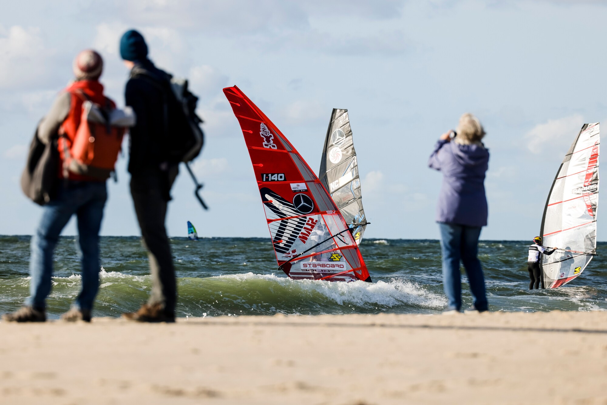 Personen am Strand beobachten Windsurfer bei einer Regatta im Meer