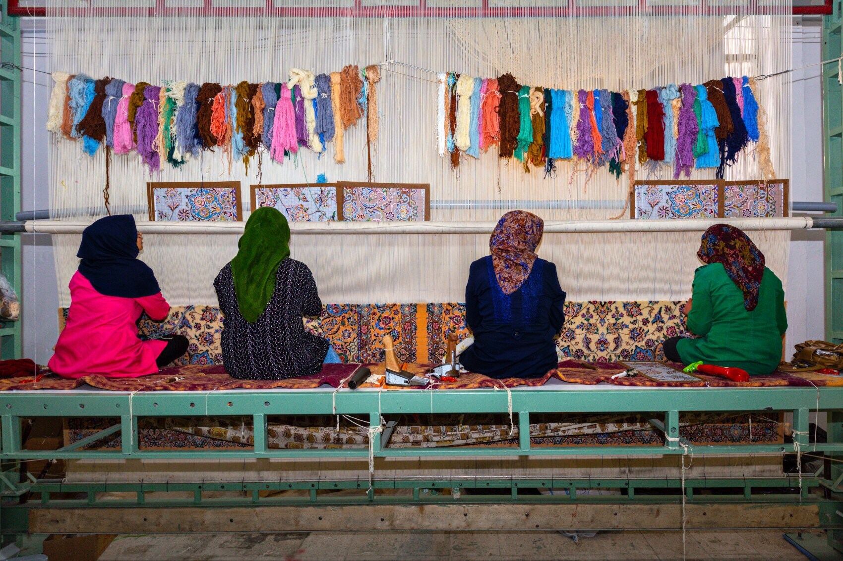 Vier Frauen sitzen mit dem Rücken zur Kamera an einem Knüpfstuhl und knüpfen einen Perserteppich