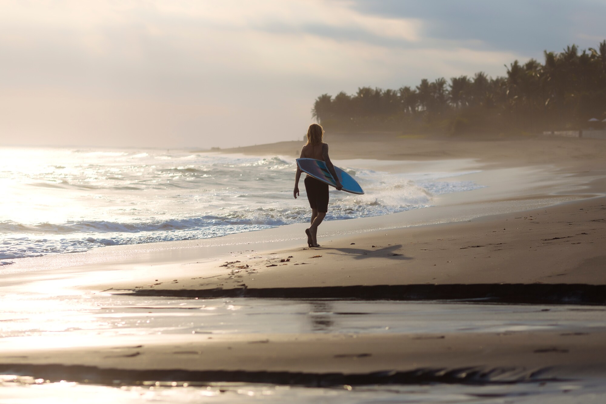 Rückansicht einer Person mit Surfbrett unter dem Arm, die einen palmengesäumten Sandstrand in der Abendsonne entlangläuft