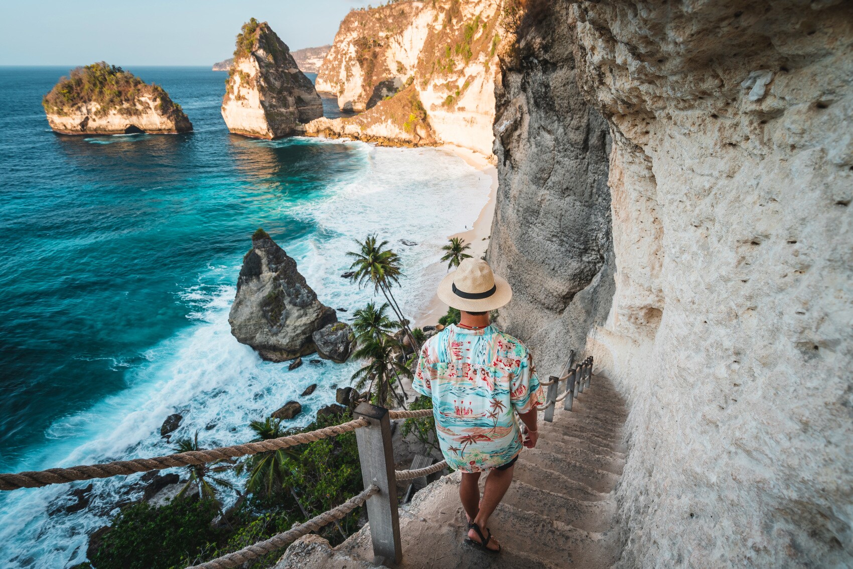 Rückansicht eines Mannes mit Strohhut und Hawaiihemd, der eine schmale Steintreppe an einer Felswand hinunter zu einer palmengesäumten Strandbucht geht Rückansicht eines Mannes mit Strohhut und Hawaiihemd, der eine schmale Steintreppe an einer Felswand hinunter zu einer palmengesäumten Strandbucht geht