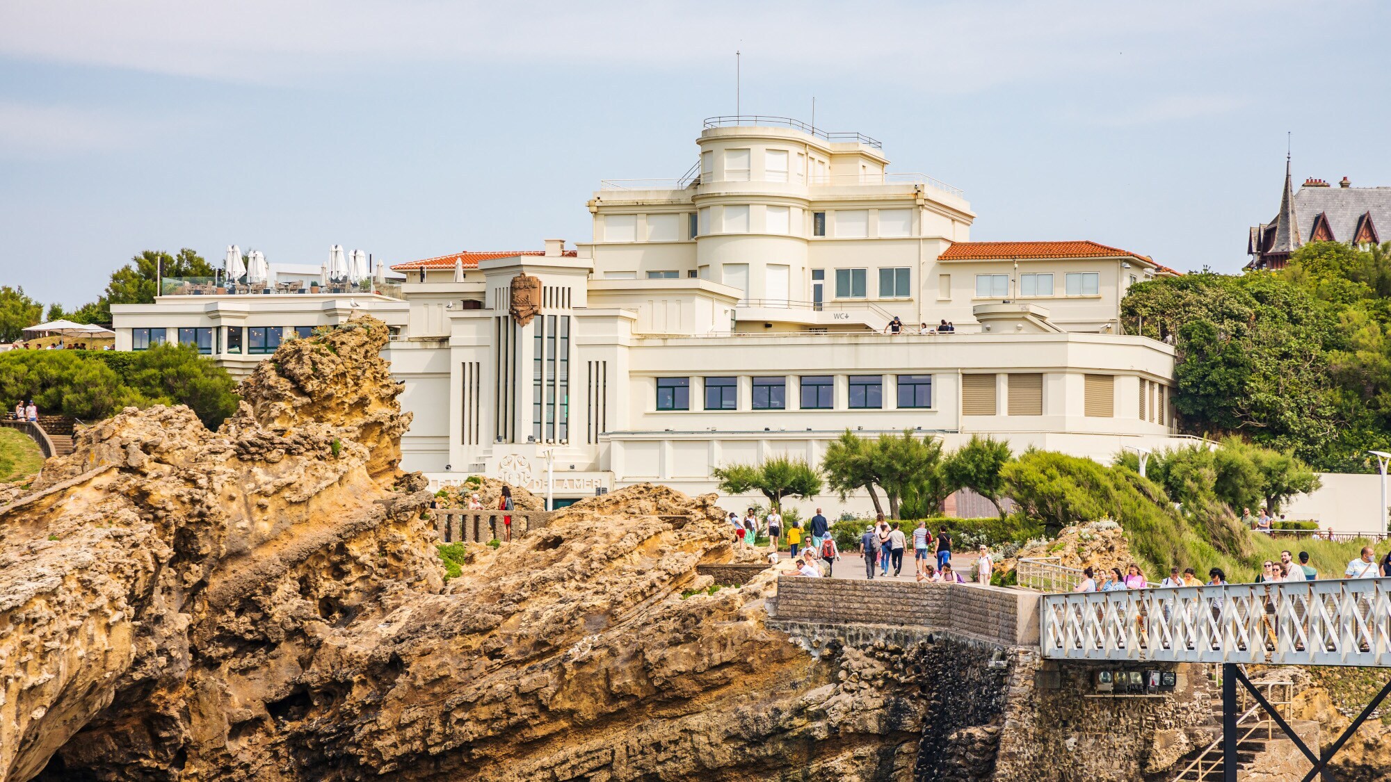 Ein helles Museumsgebäude im Art-Déco-Stil an einer Felsküste; davor Personen, die über eine Brücke laufen
