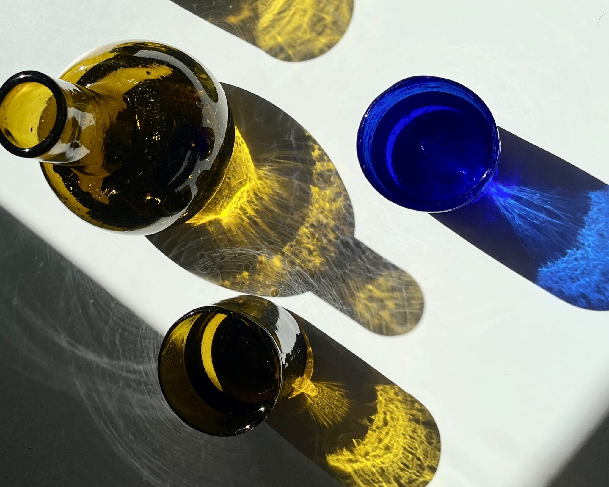 Eine olivgrüne Glaskaraffe, ein olivgrünes und ein blaues Glas, durch die von einer Seite fällt Licht fällt, wodurch farbliche Schatten entstehen.