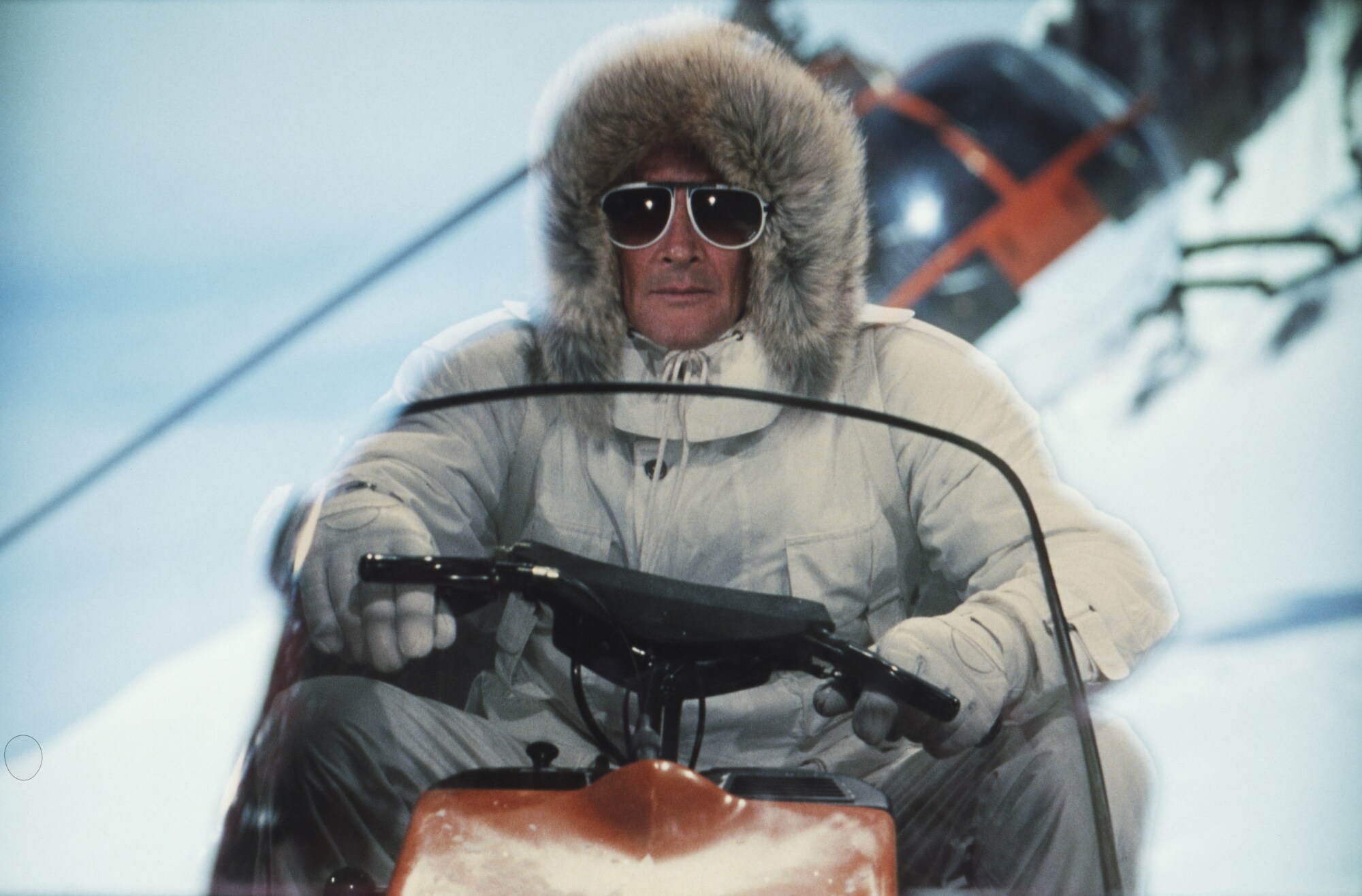 Schauspieler Roger Moore fährt im weißen Schneeanzug und großer Sonnenbrille auf einem Schneemobil
