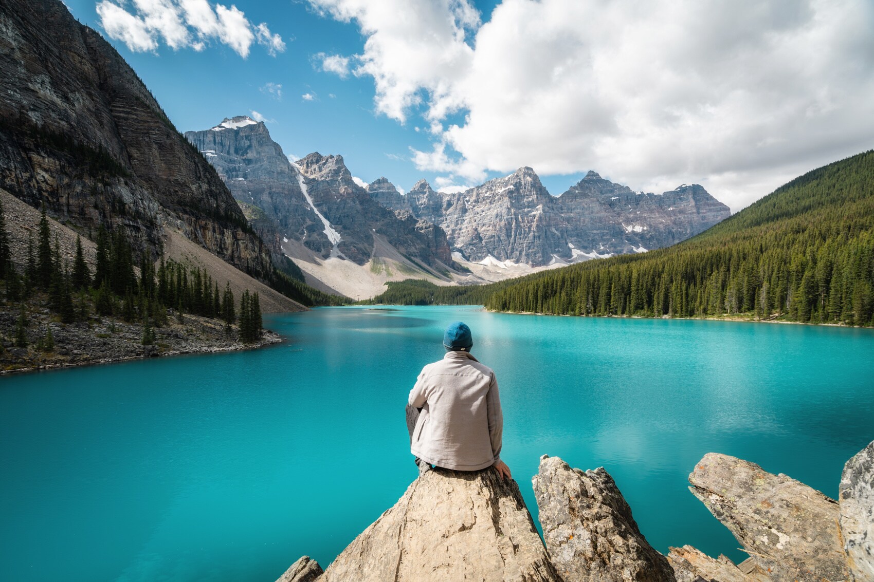 Eine Person sitzt auf einem Stein und schaut auf einen See, der gesäumt von bewaldeten Bergen ist Eine Person sitzt auf einem Stein und schaut auf einen See, der gesäumt von bewaldeten Bergen ist