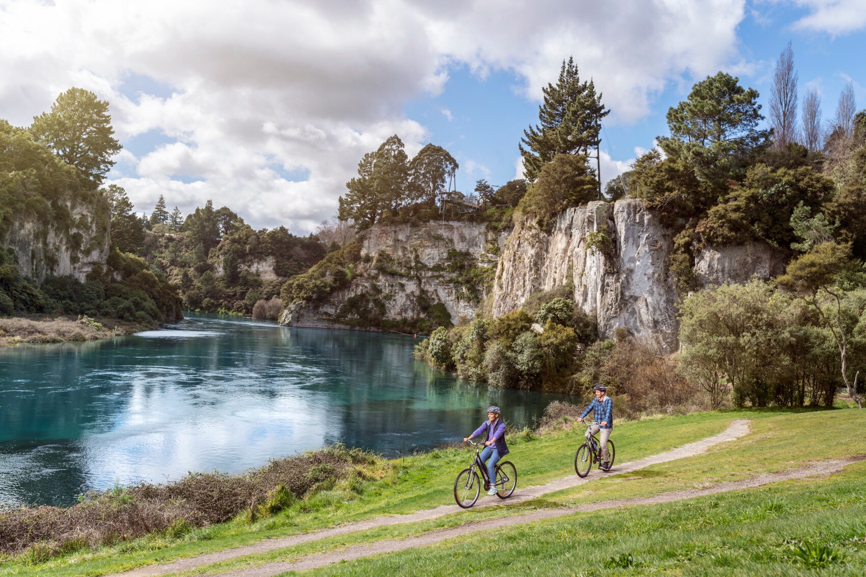 Ein Paar auf Fahrrädern fährt auf einem Radweg entlang eines Sees, umgeben von bewaldeten Felsen Ein Paar auf Fahrrädern fährt auf einem Radweg entlang eines Sees, umgeben von bewaldeten Felsen