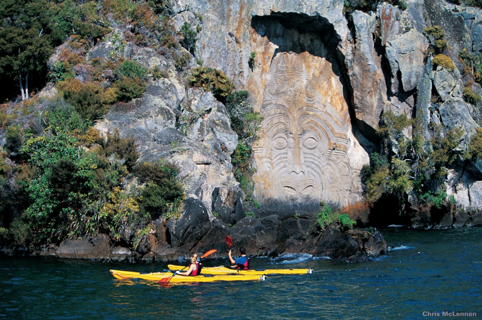 Zwei Kajakfahrer paddeln auf einem Gewässer entlang einer Felswand mit Maori-Kunst