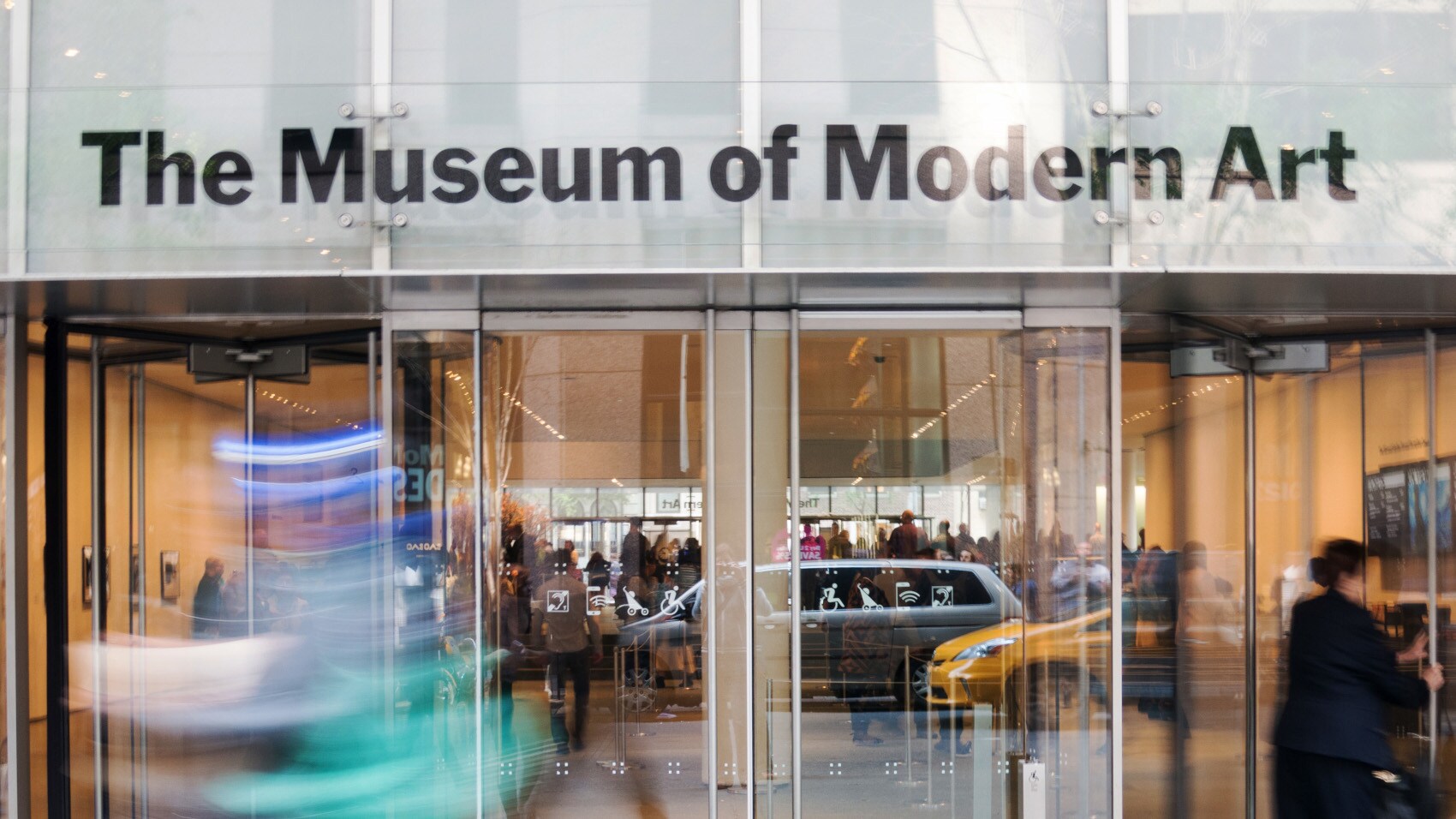 Personen am Eingang des Museum of Modern Art mit Glastüren, in denen sich auf der Straße vorbeifahrende Autos spiegeln.