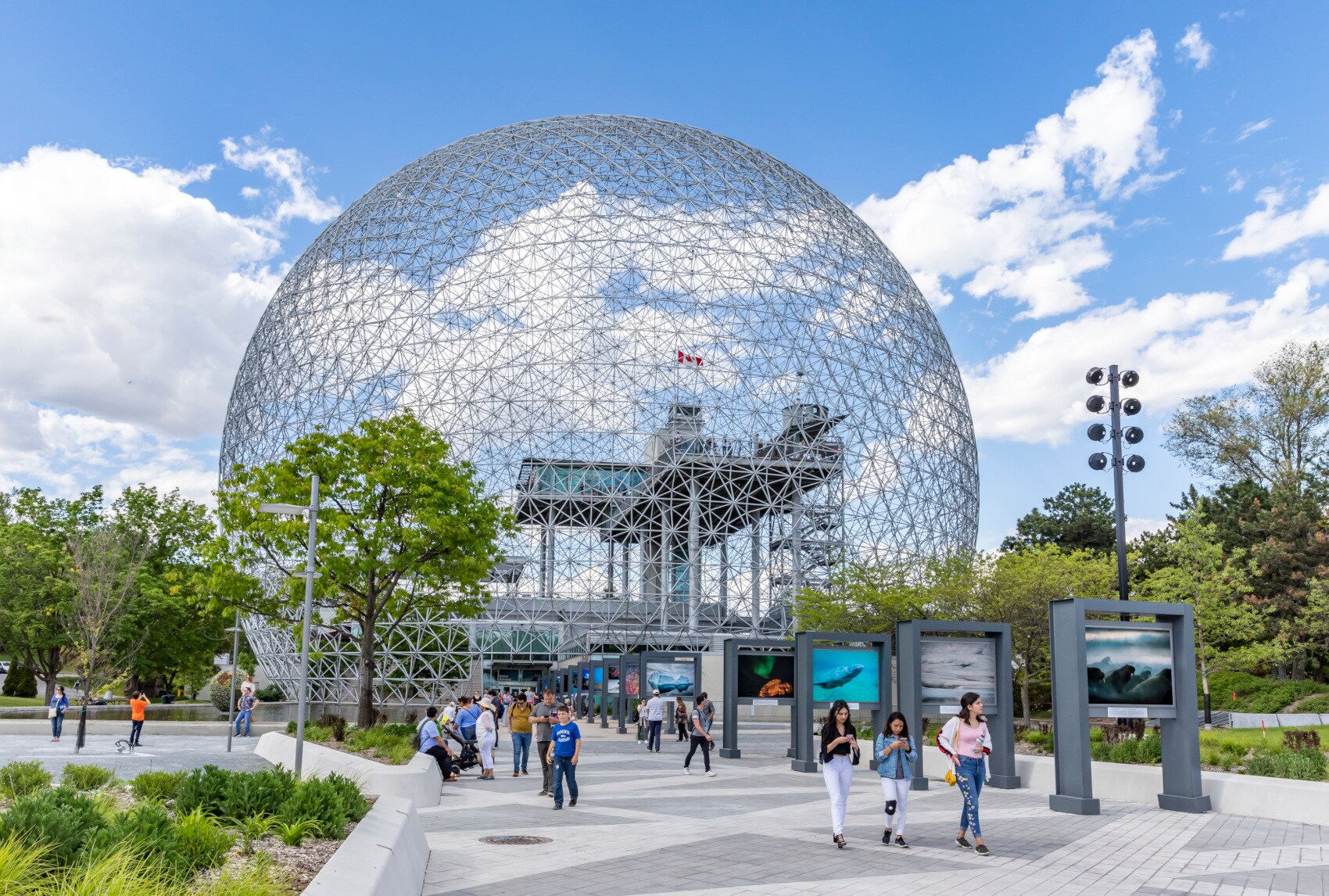 Die Stahlkuppel Biosphère in Montreal, umrahmt von Bäumen, davor Menschen Die Stahlkuppel Biosphère in Montreal, umrahmt von Bäumen, davor Menschen