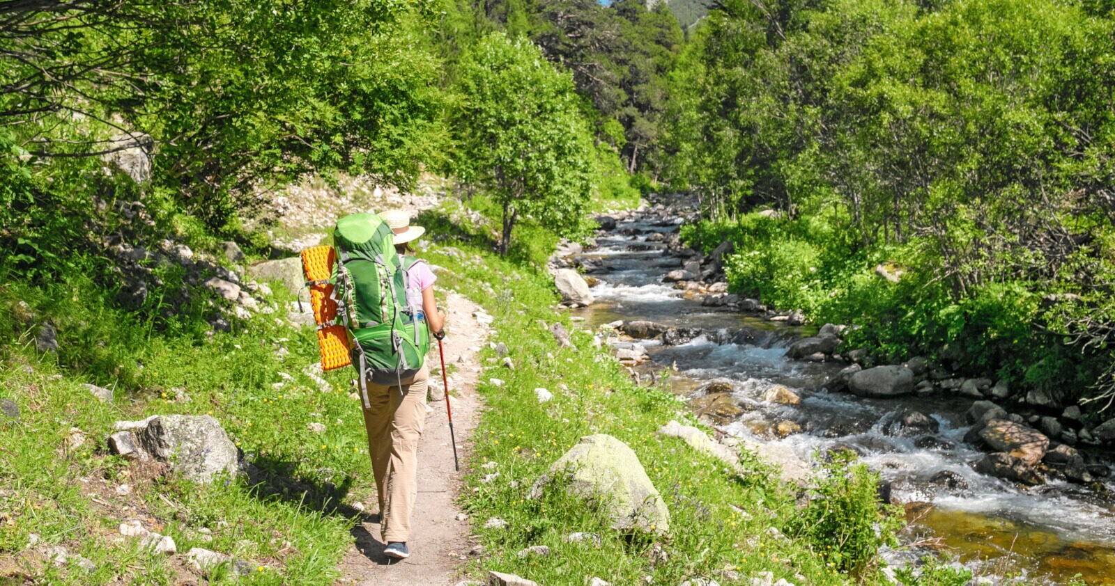 Rückansicht einer Wanderin mit Rucksack und Wanderstock, die auf einem schmalen Weg entlang eines Flusses durch eine grüne Naturlandschaft mit Bergpanorama läuft mountains Rückansicht einer Wanderin mit Rucksack und Wanderstock, die auf einem schmalen Weg entlang eines Flusses durch eine grüne Naturlandschaft mit Bergpanorama läuft