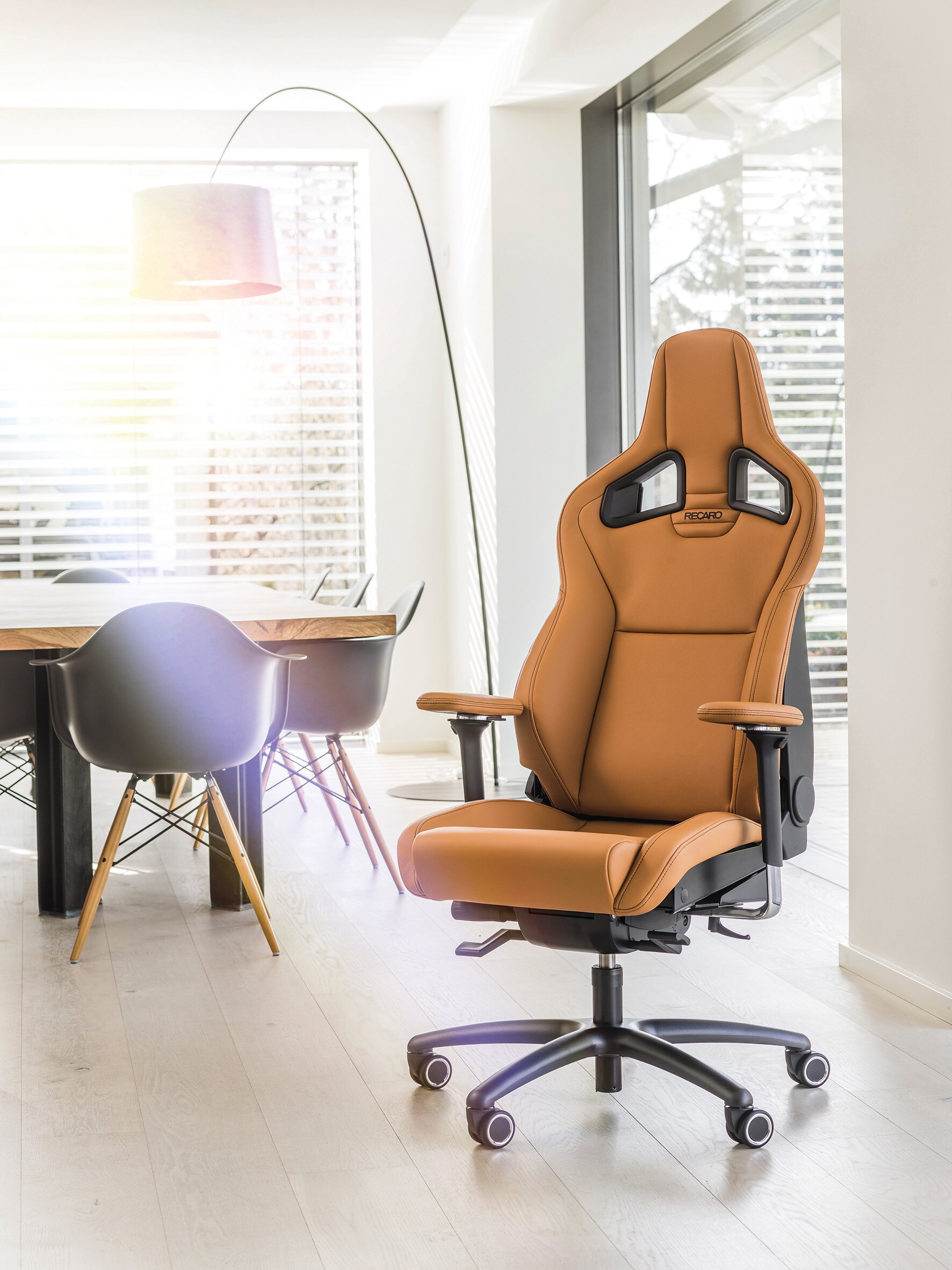 Ein cognacfarbener Bürostuhl in einem lichtdurchfluteten Raum, im Hintergrund ein Tisch mit Stühlen Ein cognacfarbener Bürostuhl in einem lichtdurchfluteten Raum, im Hintergrund ein Tisch mit Stühlen