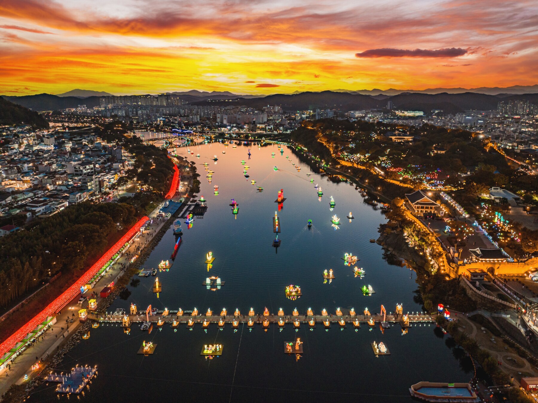 Luftaufnahme der Stadt Jinju mit einem breitem Fluss, auf dem bunte Laternen schwimmen bei Sonnenuntergang Luftaufnahme der Stadt Jinju mit einem breitem Fluss, auf dem bunte Laternen schwimmen bei Sonnenuntergang
