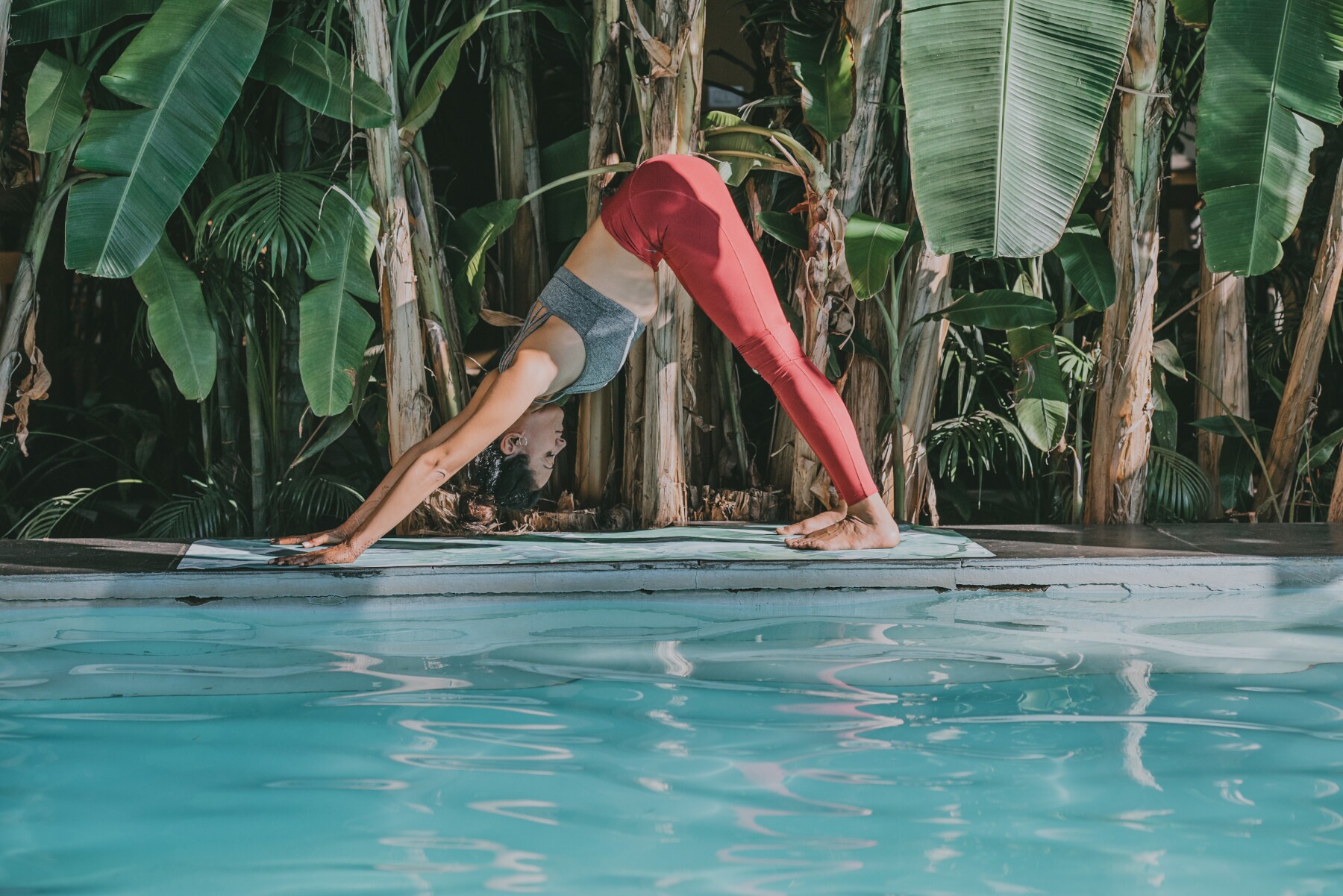 Eine junge Frau bei einer Yogaübung auf einer Matte an einem Pool, umgeben von Palmen