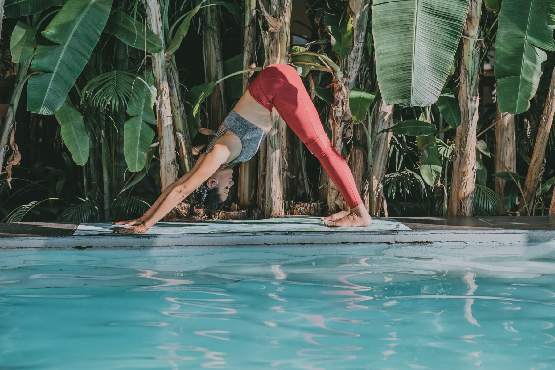 Eine junge Frau bei einer Yogaübung auf einer Matte an einem Pool, umgeben von Palmen Eine junge Frau bei einer Yogaübung auf einer Matte an einem Pool, umgeben von Palmen