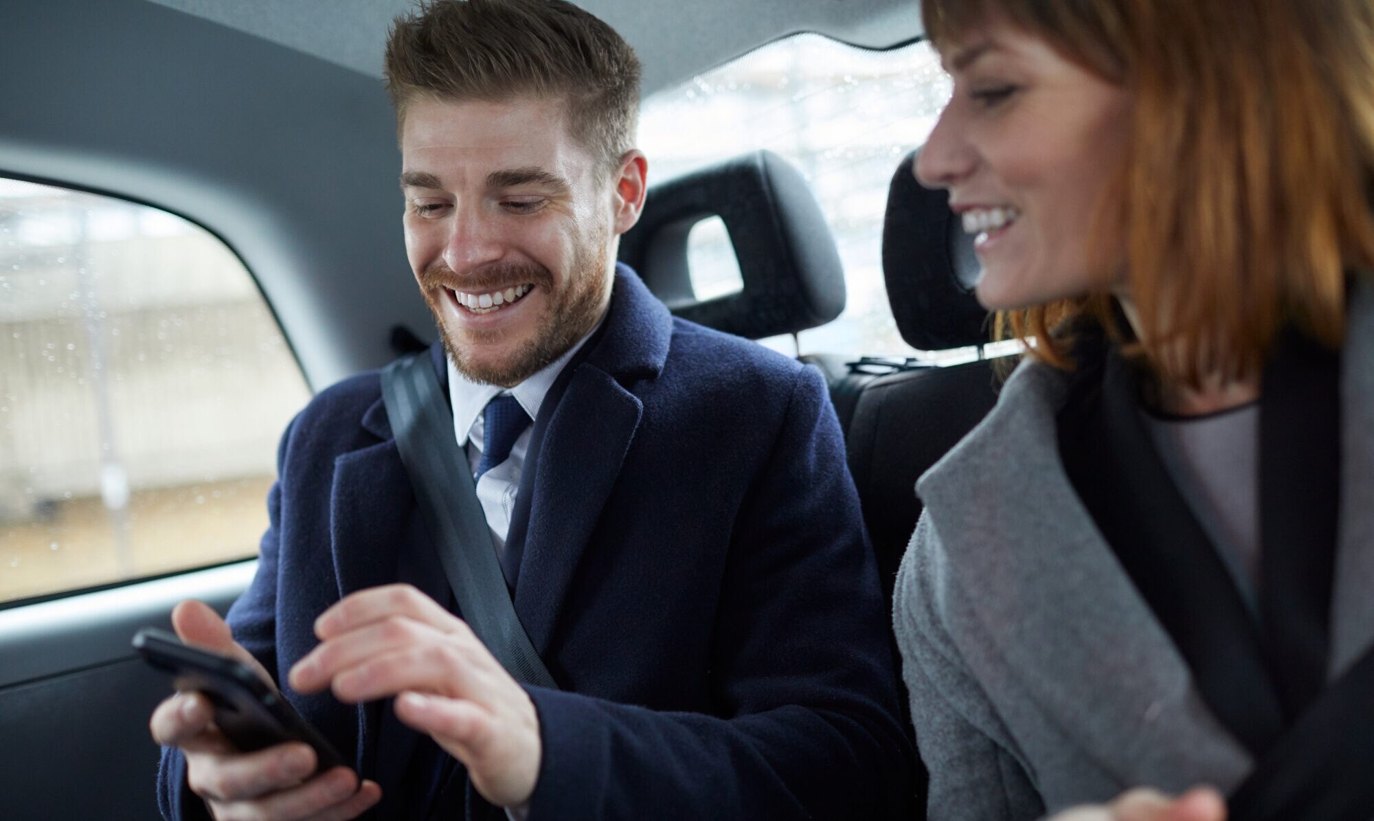Ein elegant gekleidetes Paar mittleren Alters schaut lachend auf ein Smartphone auf der Rückbank eines Autos.