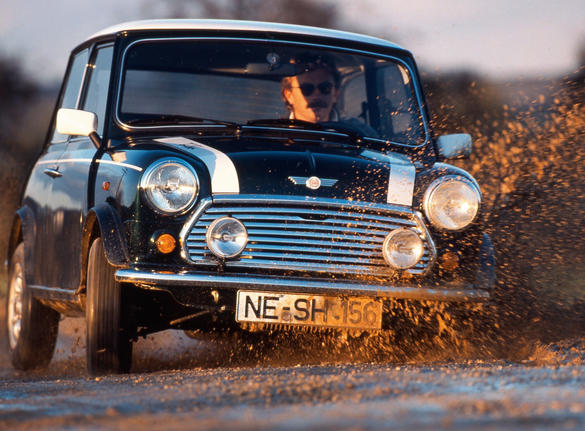 Ein Mann fährt in einem schwarzen MINI Cooper 1.3 mit Rallystreifen durch eine Schlammpfütze