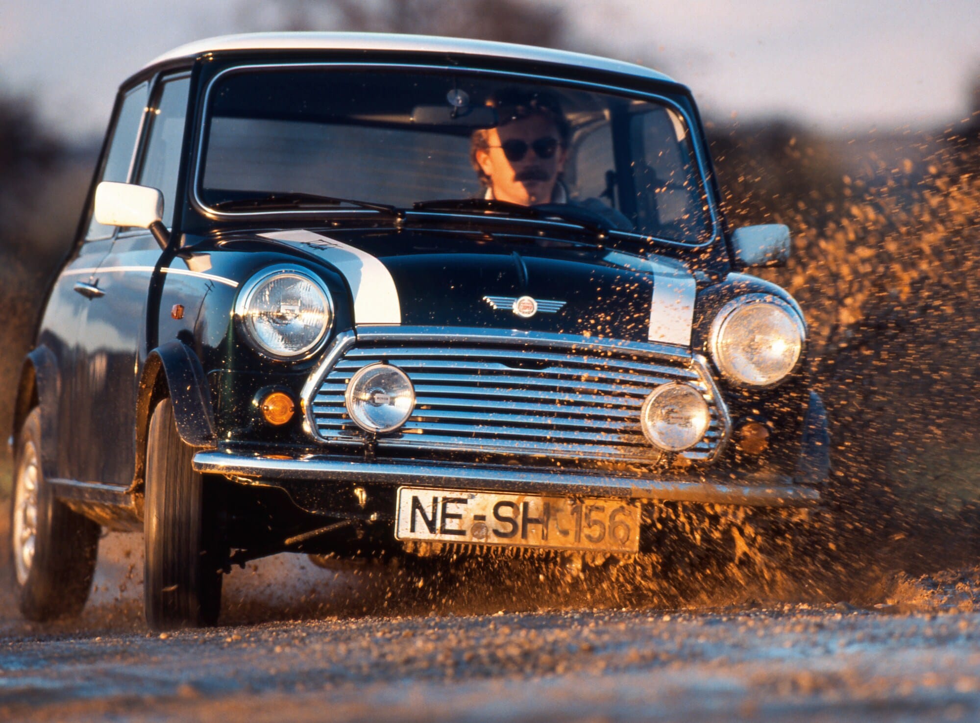 Ein Mann fährt in einem schwarzen MINI Cooper 1.3 mit Rallystreifen durch eine Schlammpfütze Ein Mann fährt in einem schwarzen MINI Cooper 1.3 mit Rallystreifen durch eine Schlammpfütze