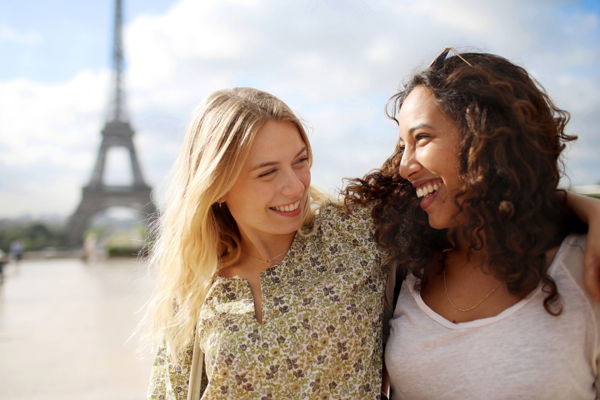 Porträt von zwei jungen Frauen, die lachend Arme umeinander legen, im Hintergrund unscharf die Silhouette des Eiffelturms. Porträt von zwei jungen Frauen, die lachend Arme umeinander legen, im Hintergrund unscharf die Silhouette des Eiffelturms.
