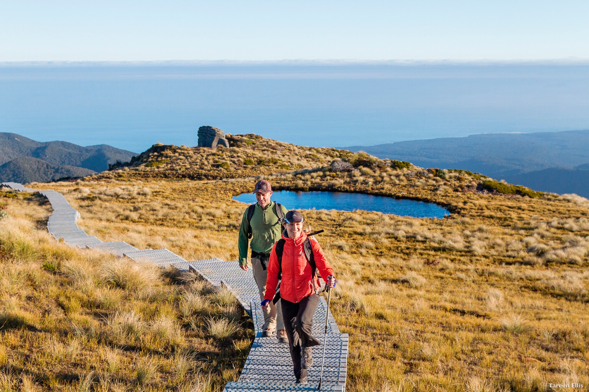 Zwei Personen in Funktionsbekleidung mit Wanderstöcken gehen auf einem Holzsteg durch eine Graslandschaft auf einem Berg.