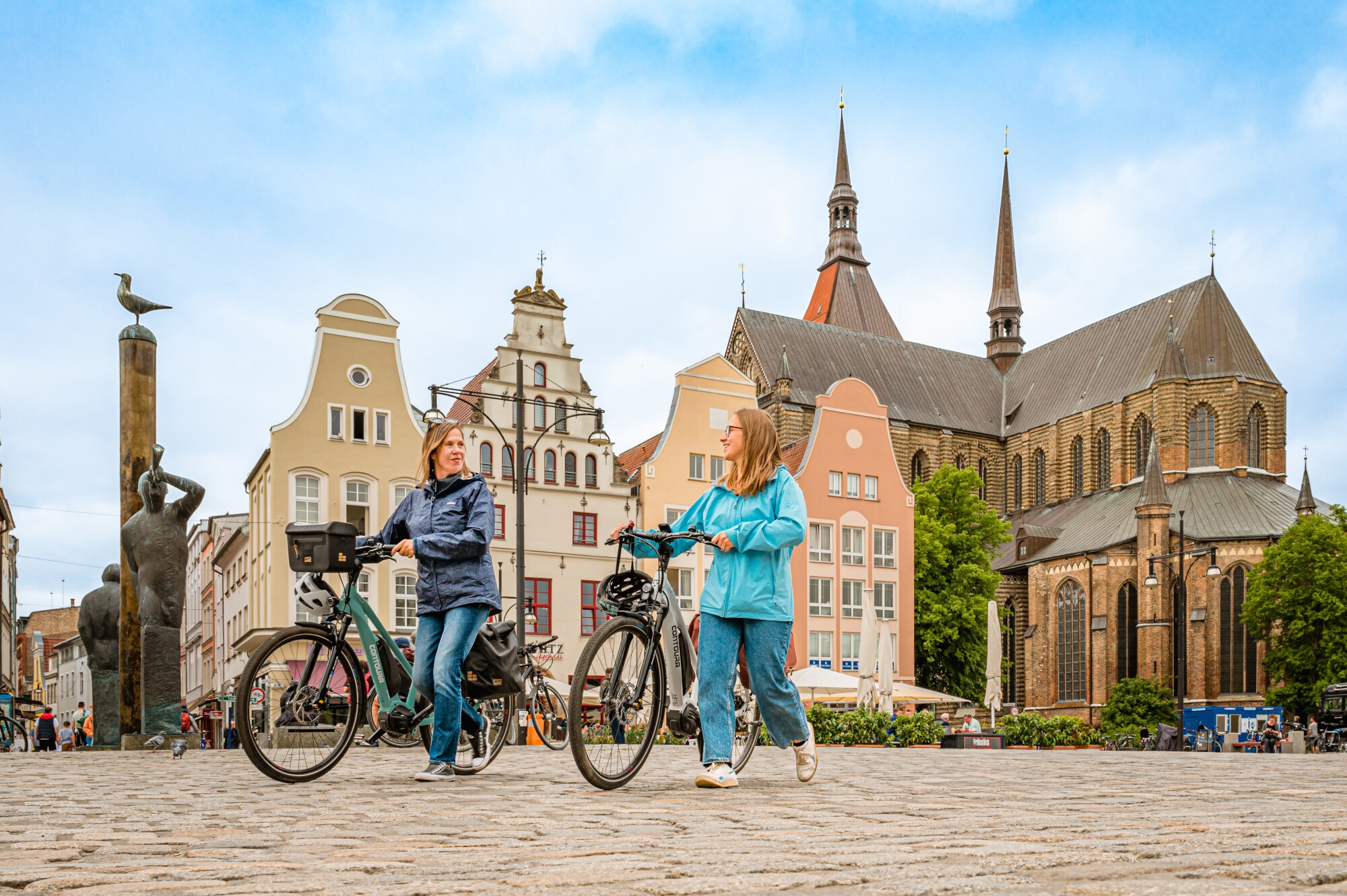 Zwei Frauen schieben nebeneinander ihre E-Bikes, im Hintergrund historische Gebäude in Rostock Zwei Frauen schieben nebeneinander ihre E-Bikes, im Hintergrund historische Gebäude in Rostock