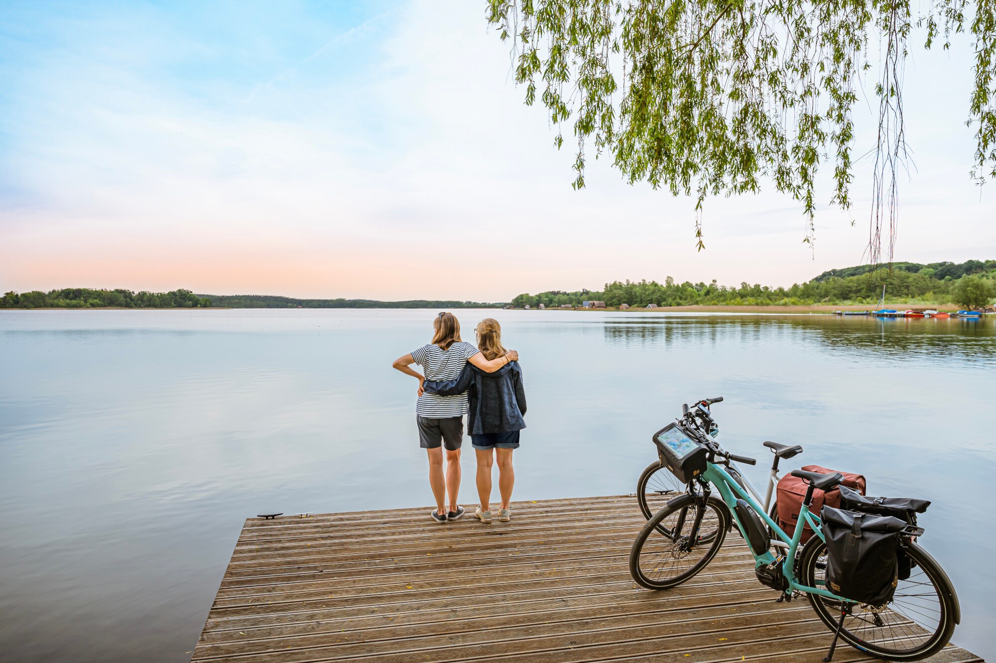 Rückansicht von zwei Frauen, die auf einem Steg vor einem See stehen und sich umarmen, daneben zwei E-Bikes Rückansicht von zwei Frauen, die auf einem Steg vor einem See stehen und sich umarmen, daneben zwei E-Bikes