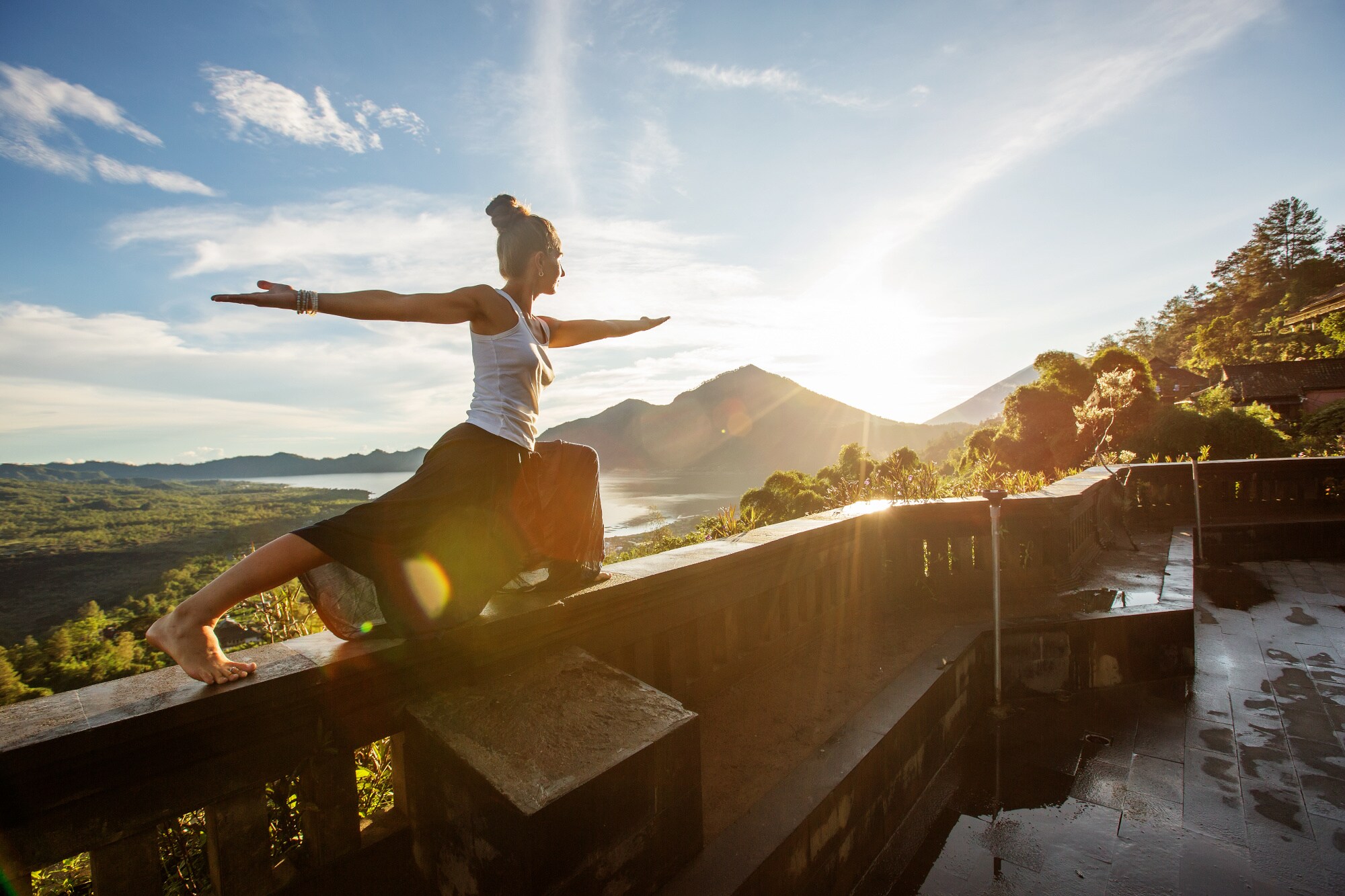 Eine Frau hält eine Yogaposition auf dem Terrassengeländer mit Blick in die weite Landschaft eines Regenwaldes bei Sonnenaufgang