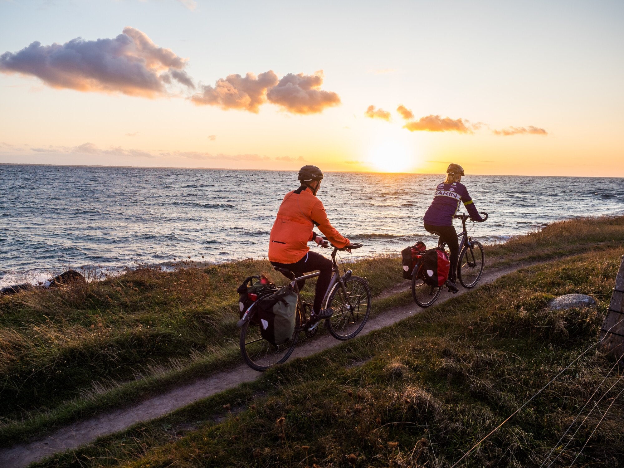 Rückansicht von zwei Personen auf Fahrrädern, die auf einem schmalen Radweg eine Küste bei Sonnenuntergang entlangfahren Rückansicht von zwei Personen auf Fahrrädern, die auf einem schmalen Radweg eine Küste bei Sonnenuntergang entlangfahren